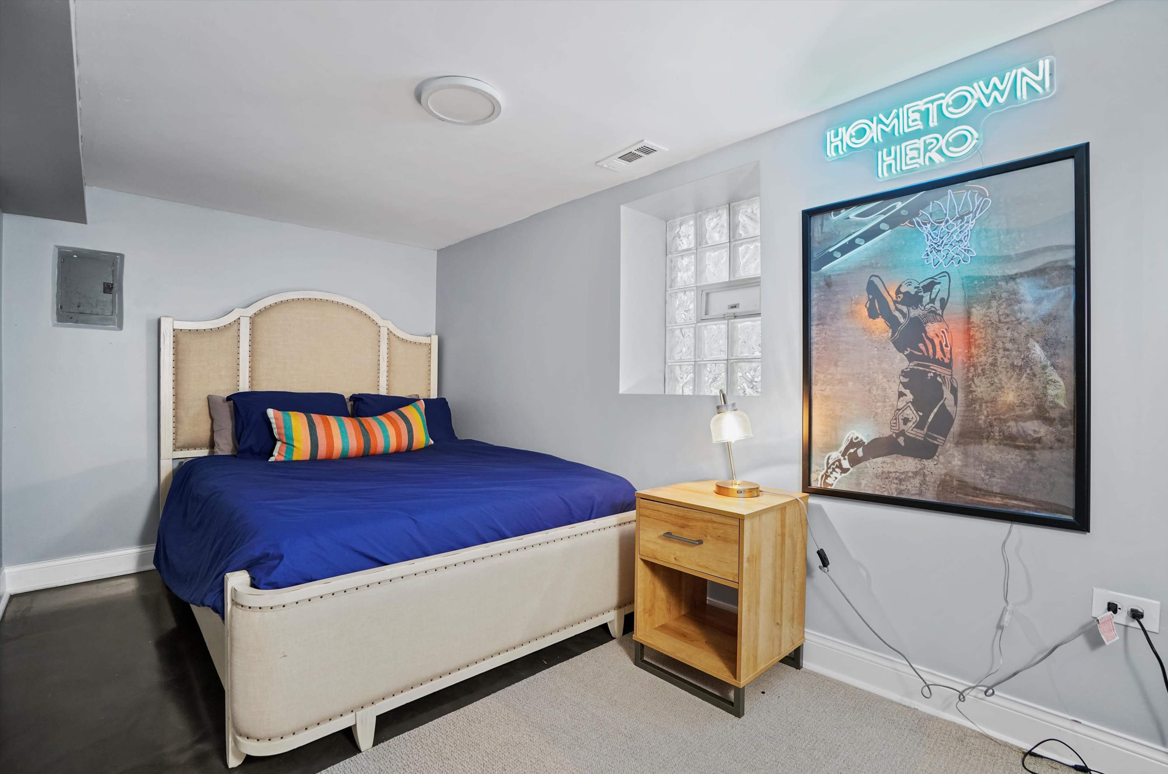 A cozy bedroom features a bed with a blue comforter, a wooden nightstand, and a neon sign on the wall.