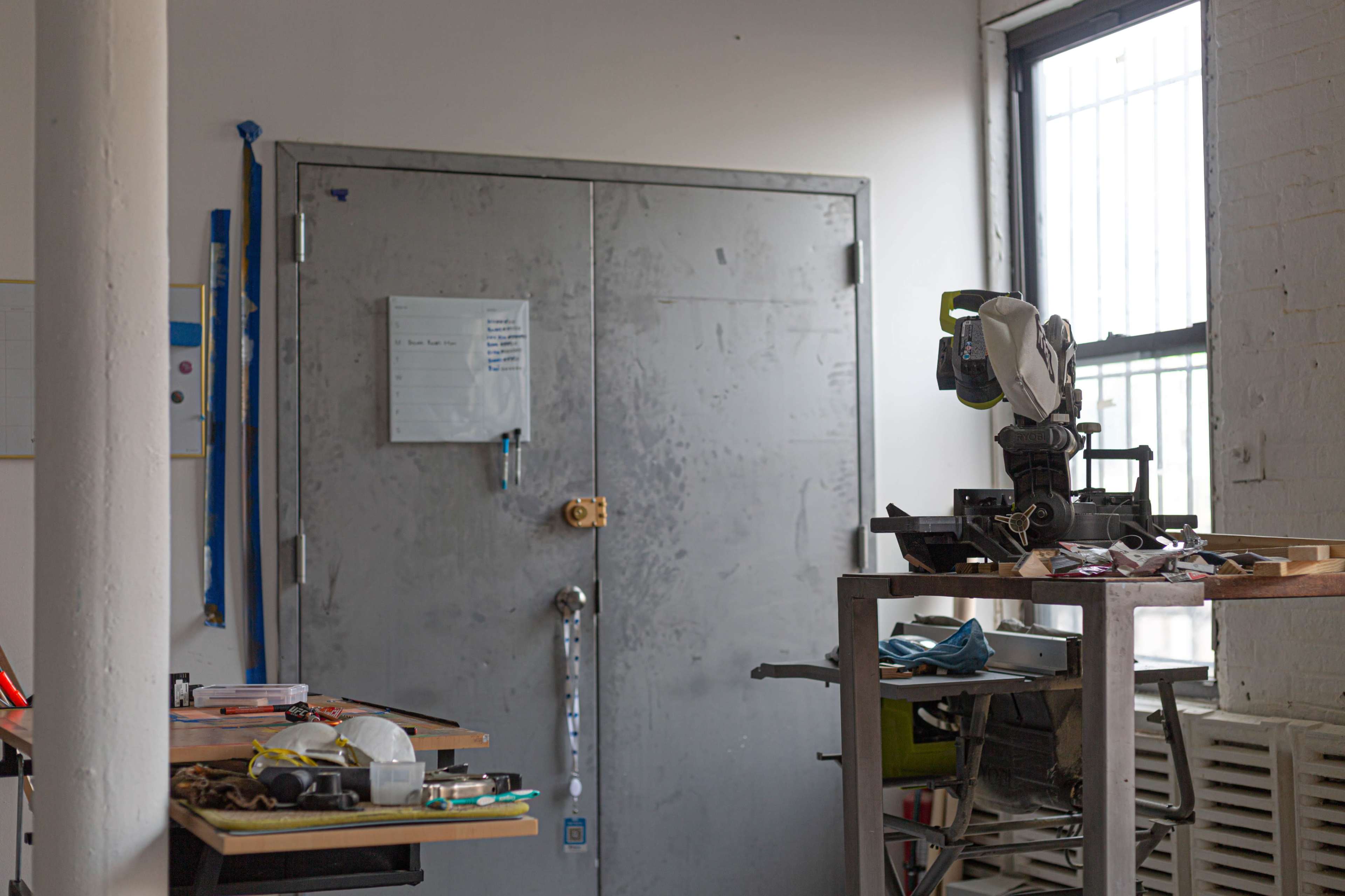 A gray metal door with a padlock is positioned at the back of a workshop, while various tools and materials are scattered on tables and a cart nearby.