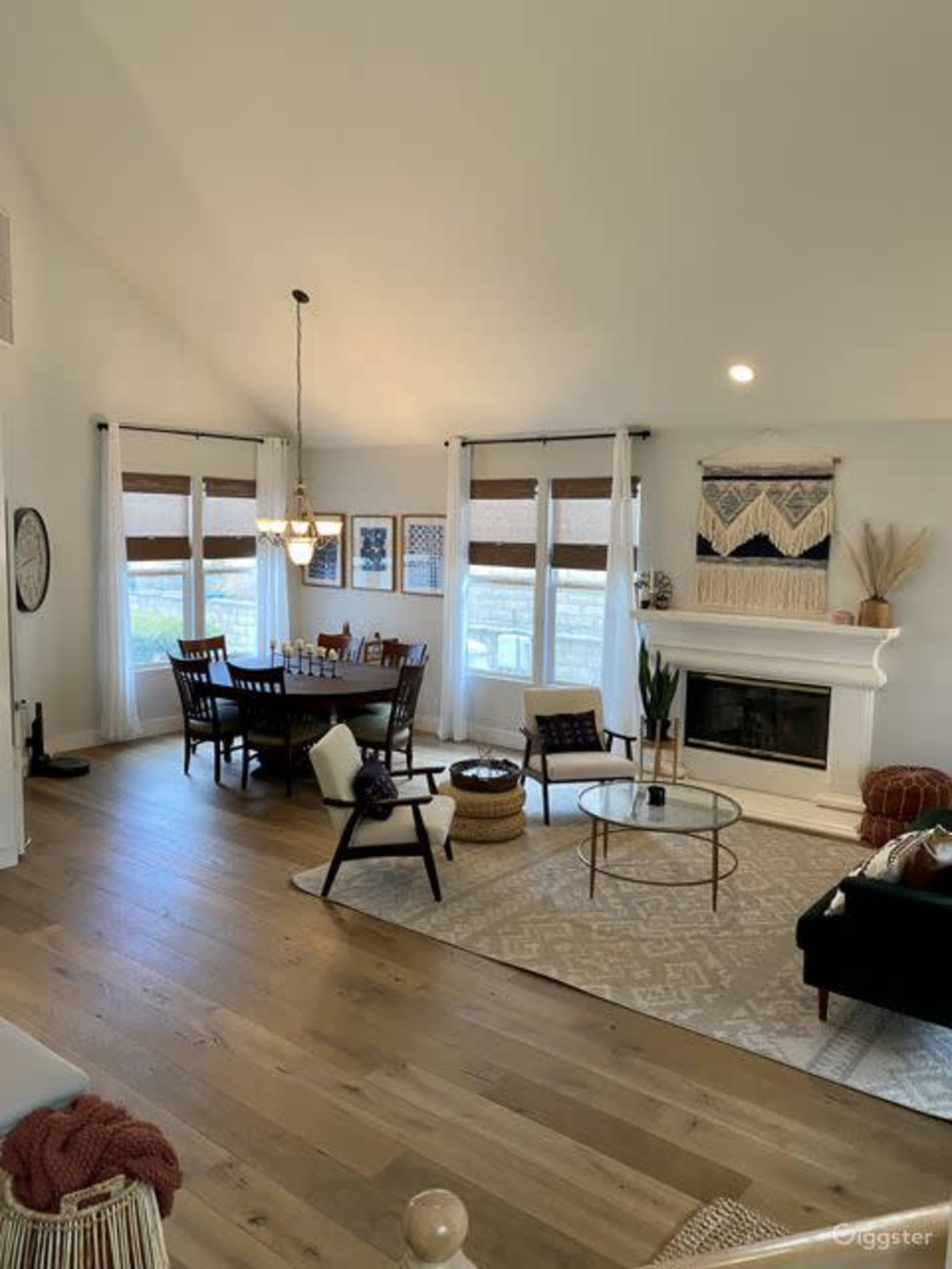 The image shows a bright living and dining area featuring a dining table, chairs, a fireplace with decor, and large windows with natural light.