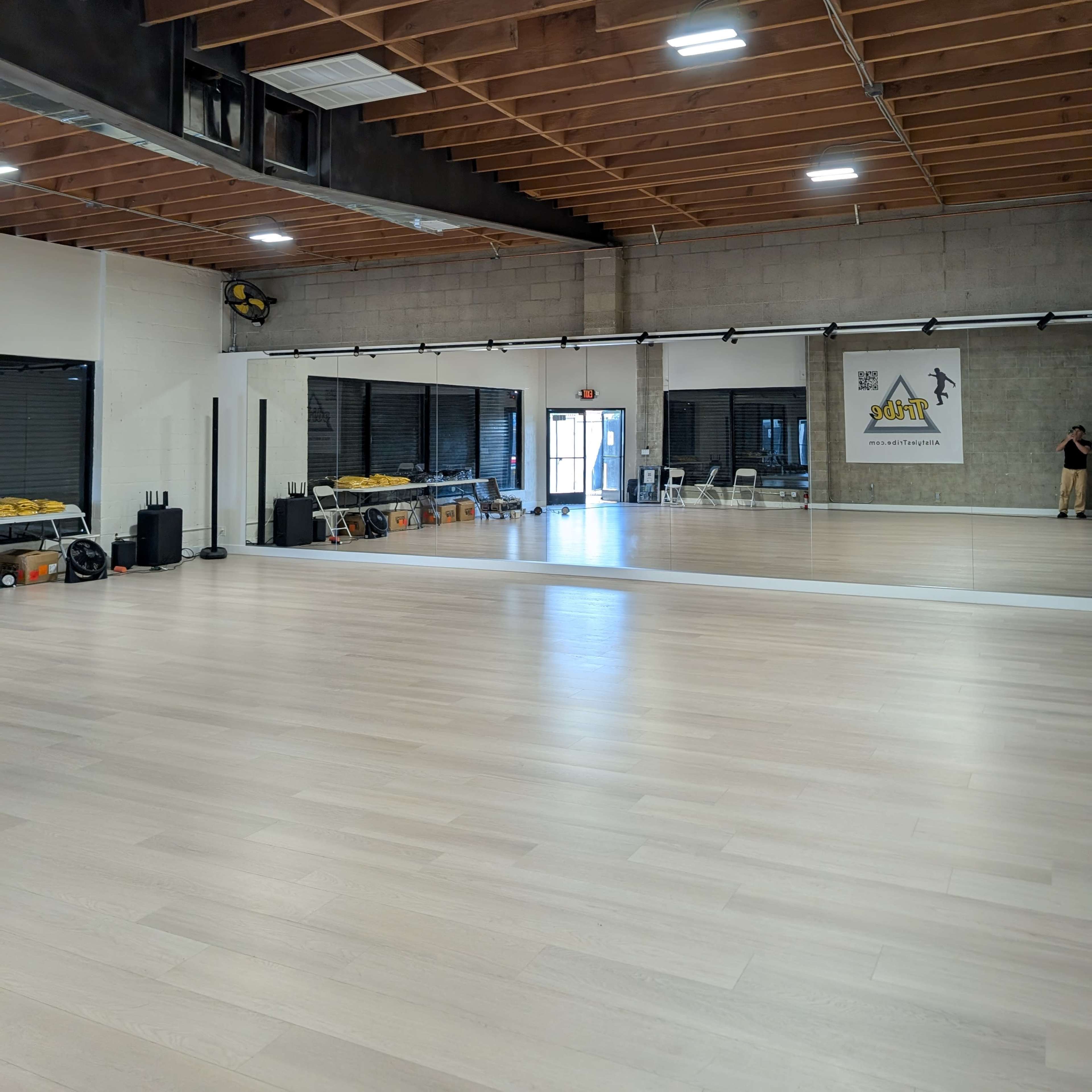 The image shows a spacious, empty dance studio featuring a mirrored wall, polished wooden floors, and large windows letting in natural light.