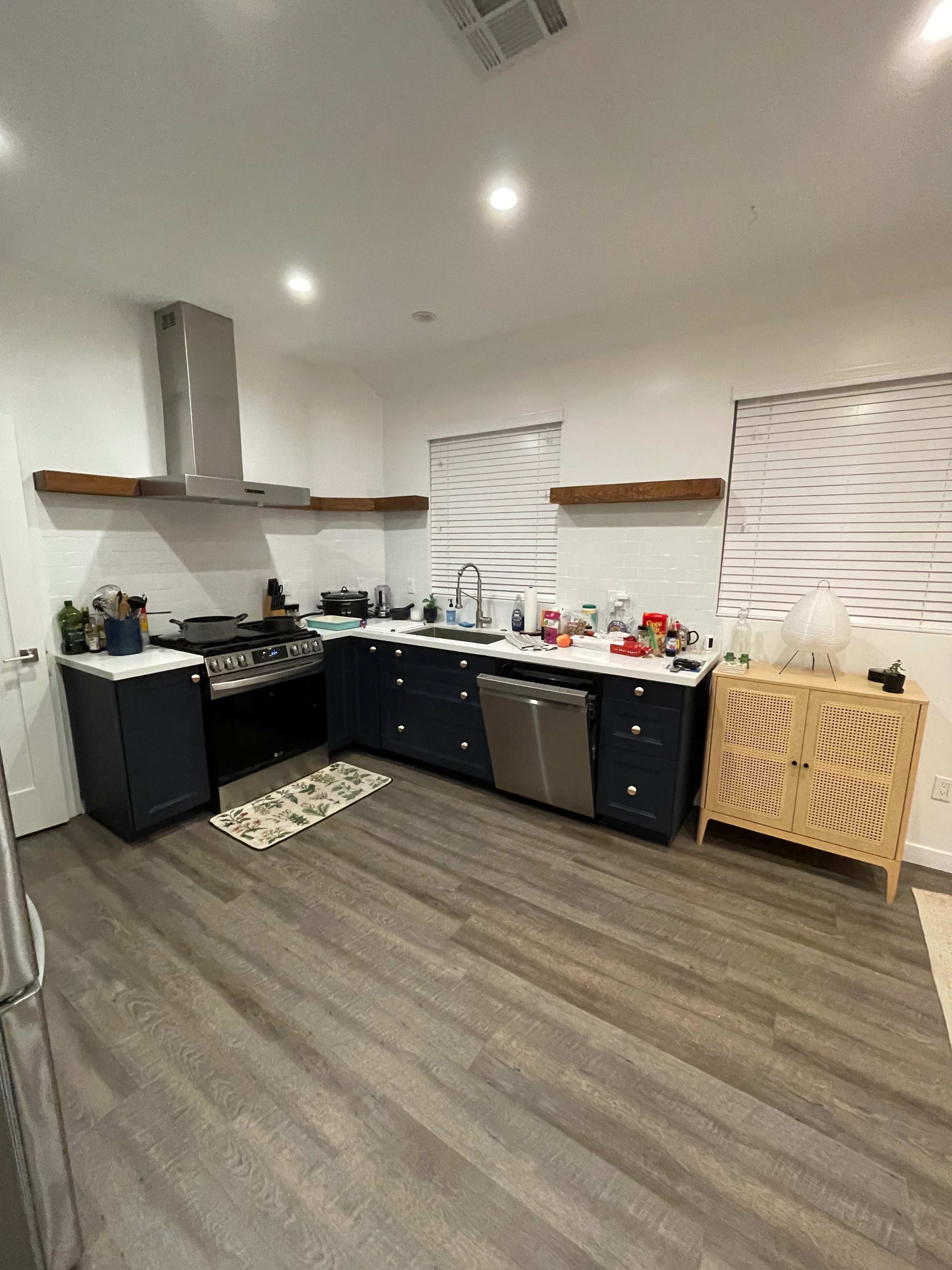 The image shows a modern kitchen featuring dark blue cabinetry, a stainless steel stove, and open shelving, with various kitchen items and utensils scattered on the countertops.