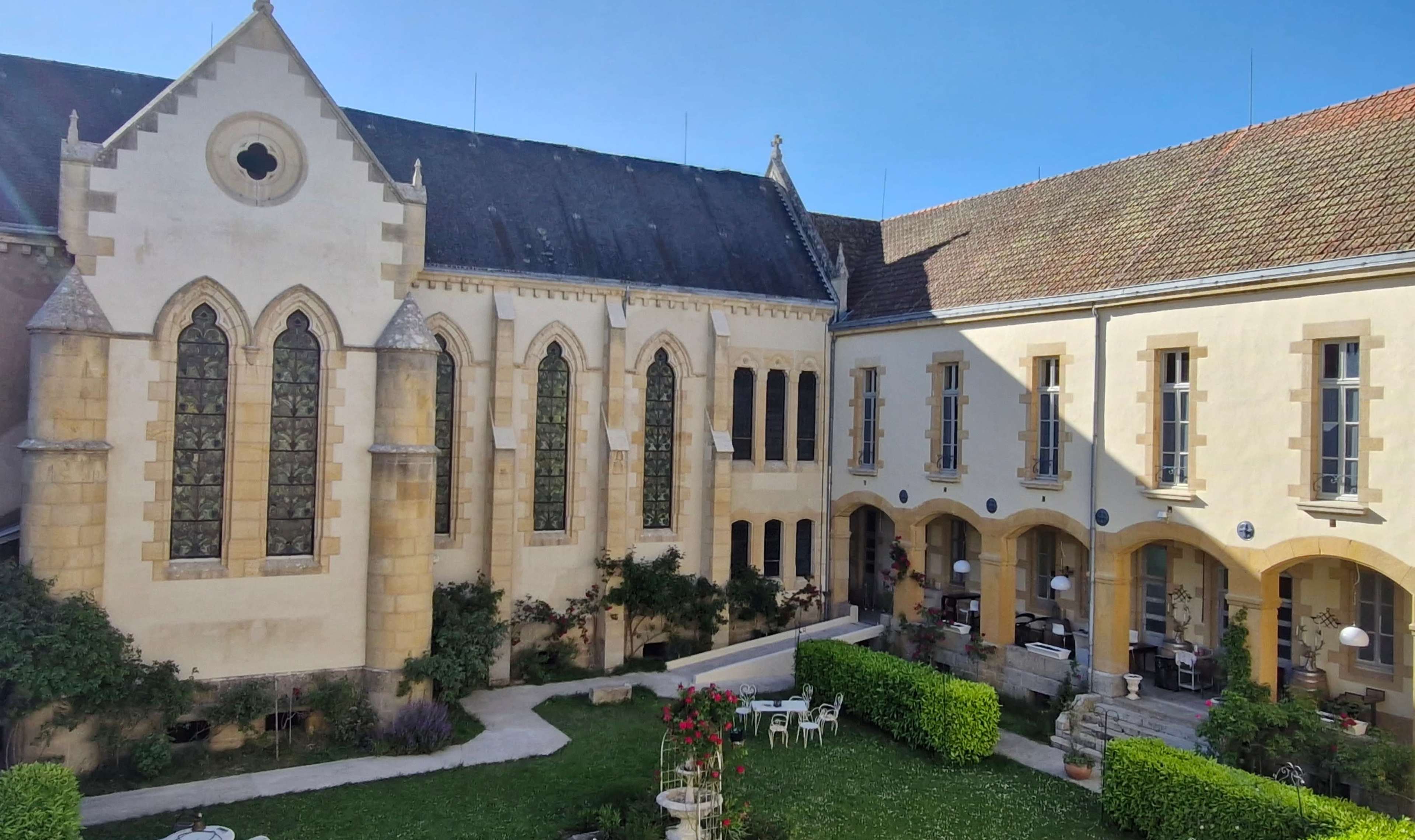 The image shows a courtyard surrounded by two buildings, one with pointed arches and large windows, and the other featuring arched doorways and a manicured green space with outdoor seating.