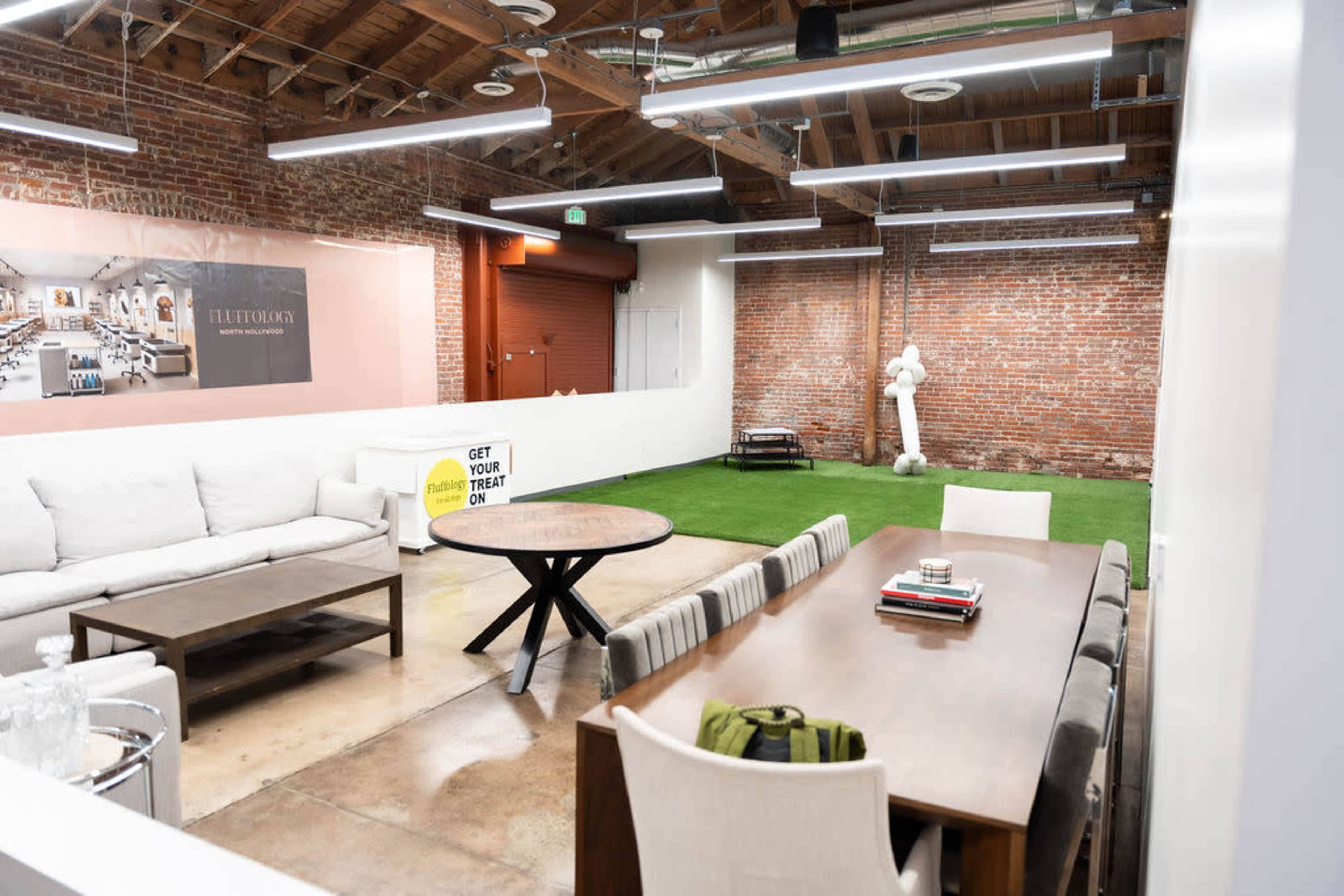 The image shows a modern workspace with exposed brick walls, a seating area featuring a large couch and a table, and an artificial grass area with a decorative white sculpture.