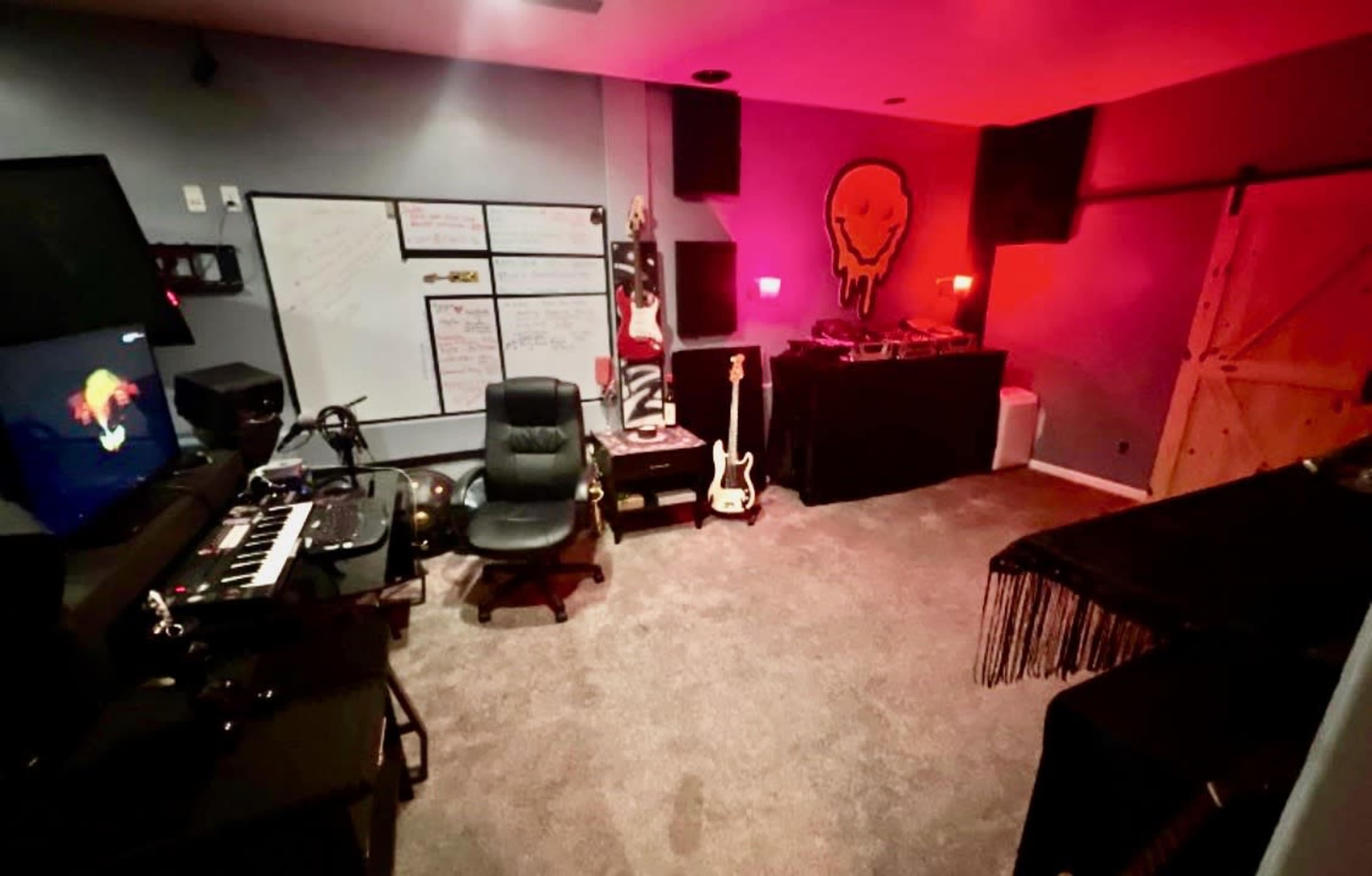 The image shows a music studio with a keyboard, guitar, and sound equipment in a dimly lit room, featuring a large wall-mounted screen and a whiteboard.