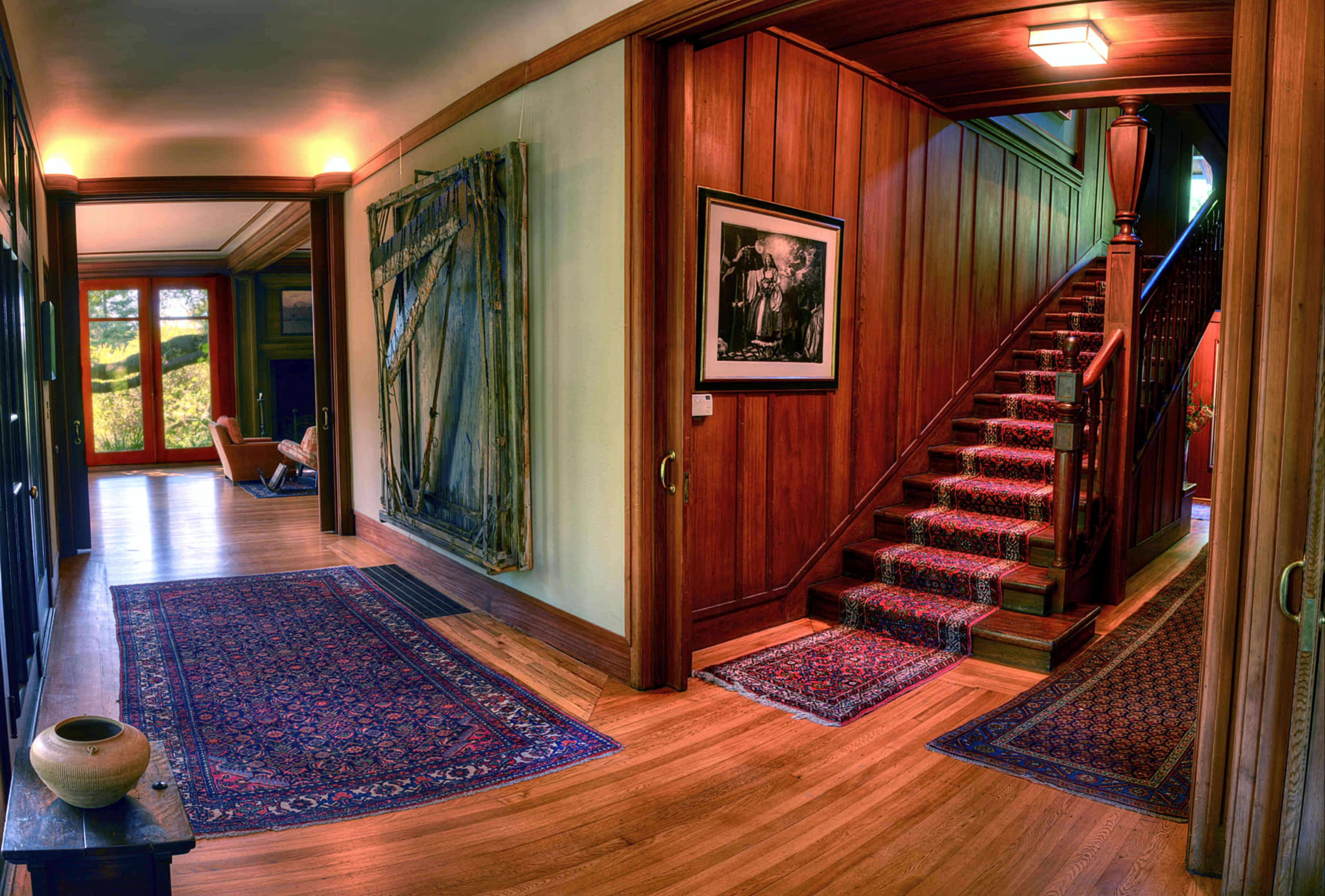 A foyer features wooden walls, a staircase with a patterned runner, and a large painting on the wall, flanked by rugs and a decorative vase.