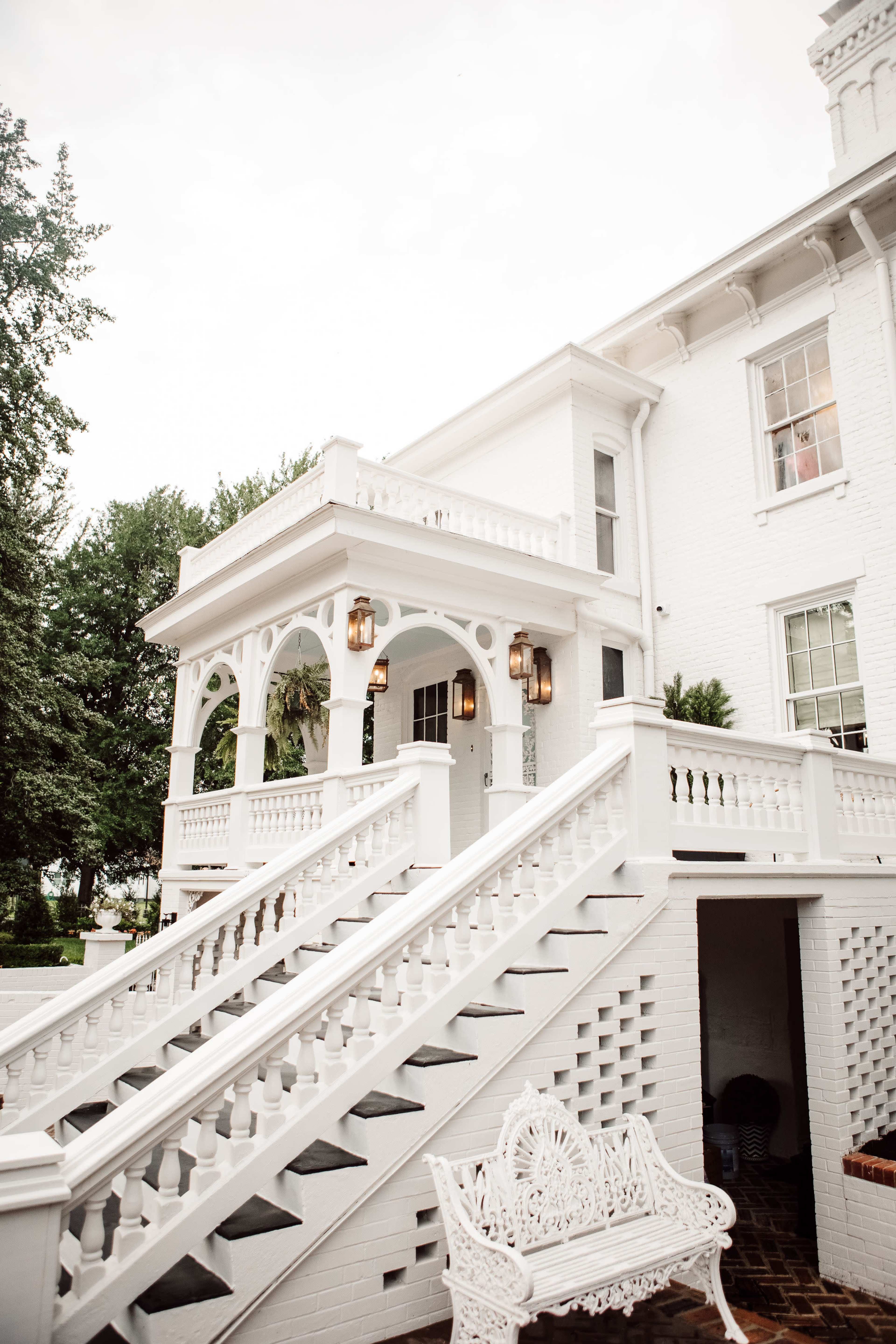 The image shows a large white house with a staircase leading up to a covered porch, and decorative details on the railing and lamps.