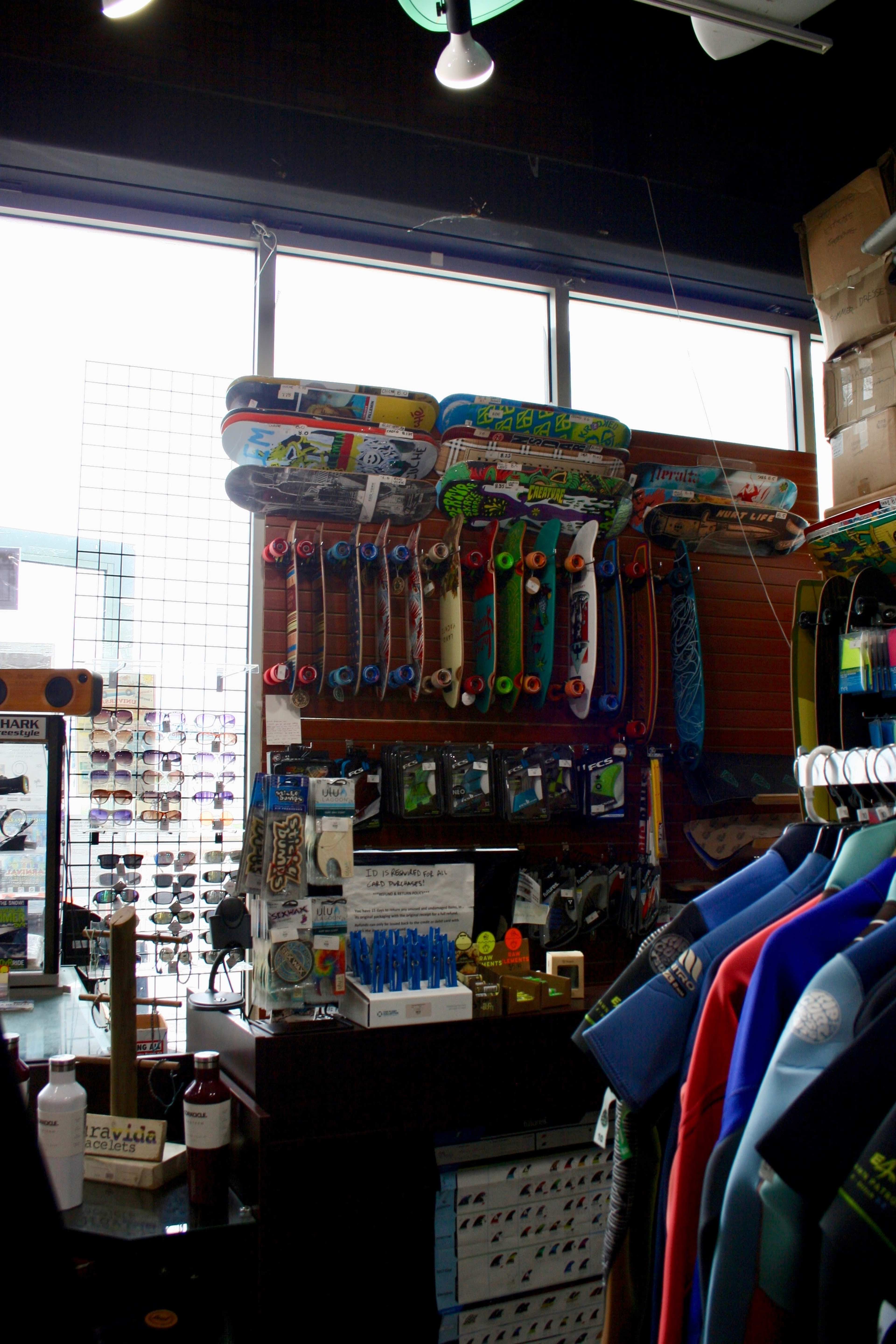 The image shows a skateboard shop interior with various skateboards displayed on a wall and merchandise, including sunglasses and wetsuits, visible throughout the store.