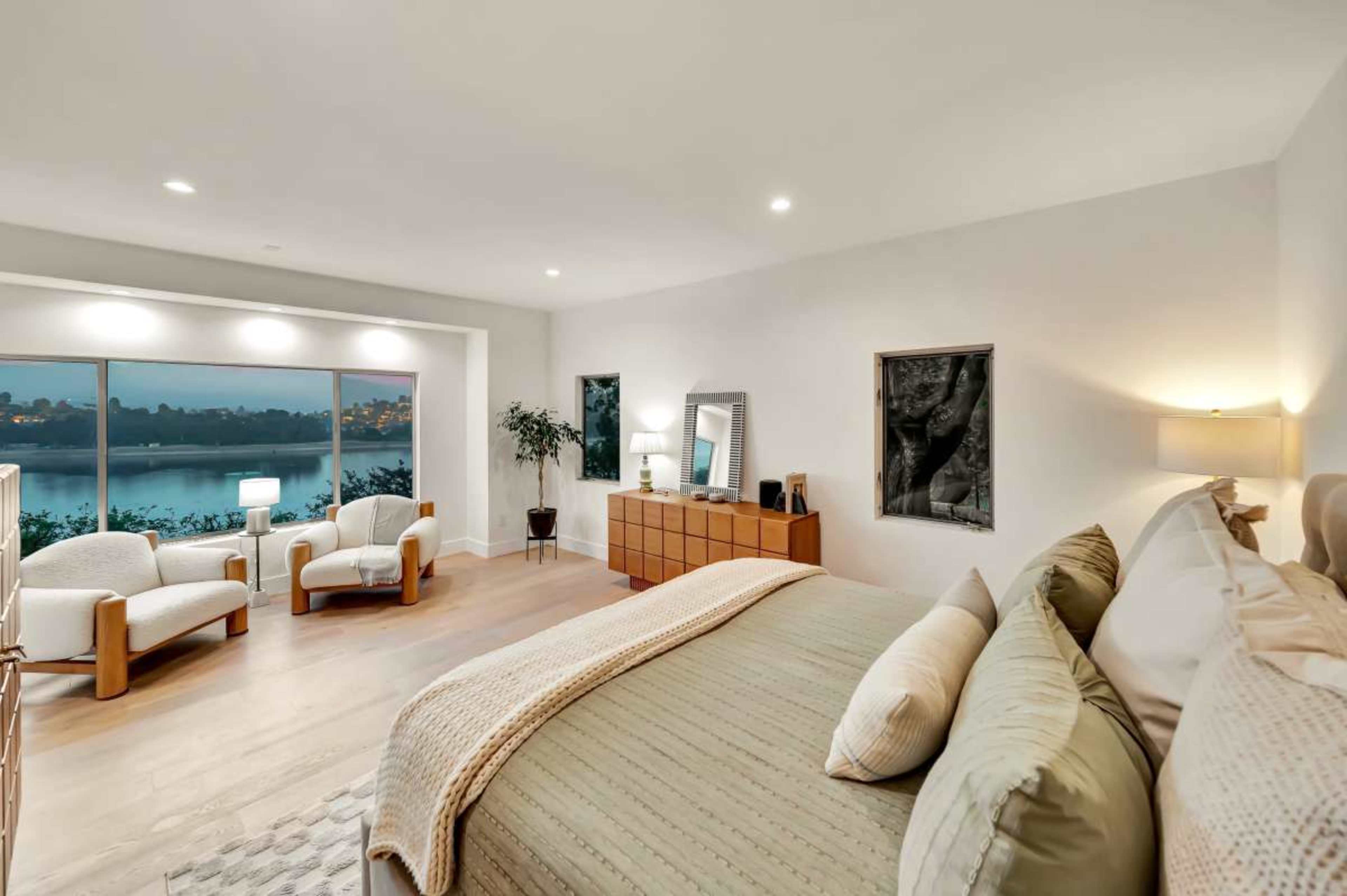 The image depicts a modern bedroom with a large window overlooking a body of water, featuring a bed, a wooden dresser, and two armchairs.