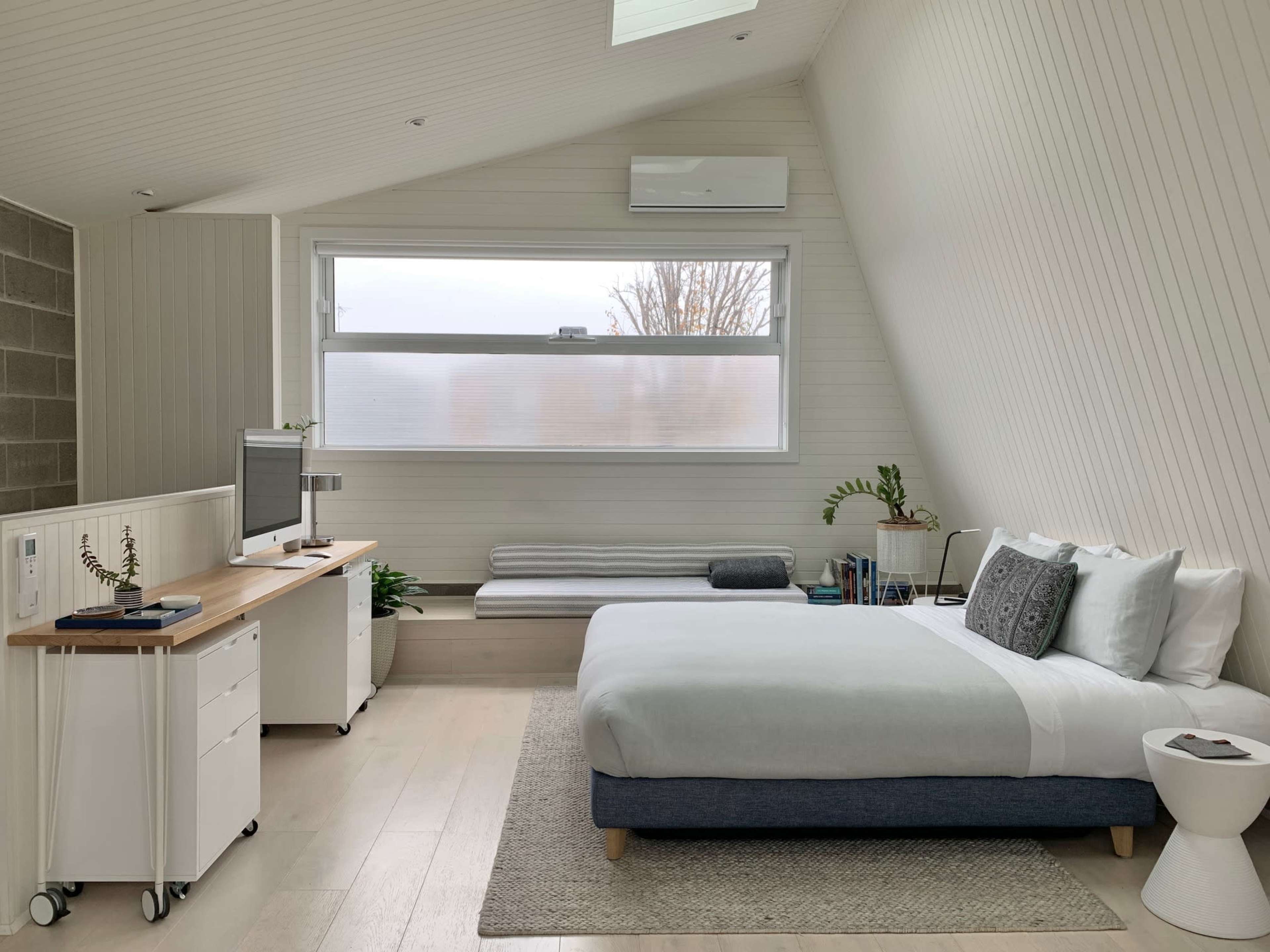 A modern bedroom with a large bed, a desk with a computer, and a window allowing natural light to fill the space.