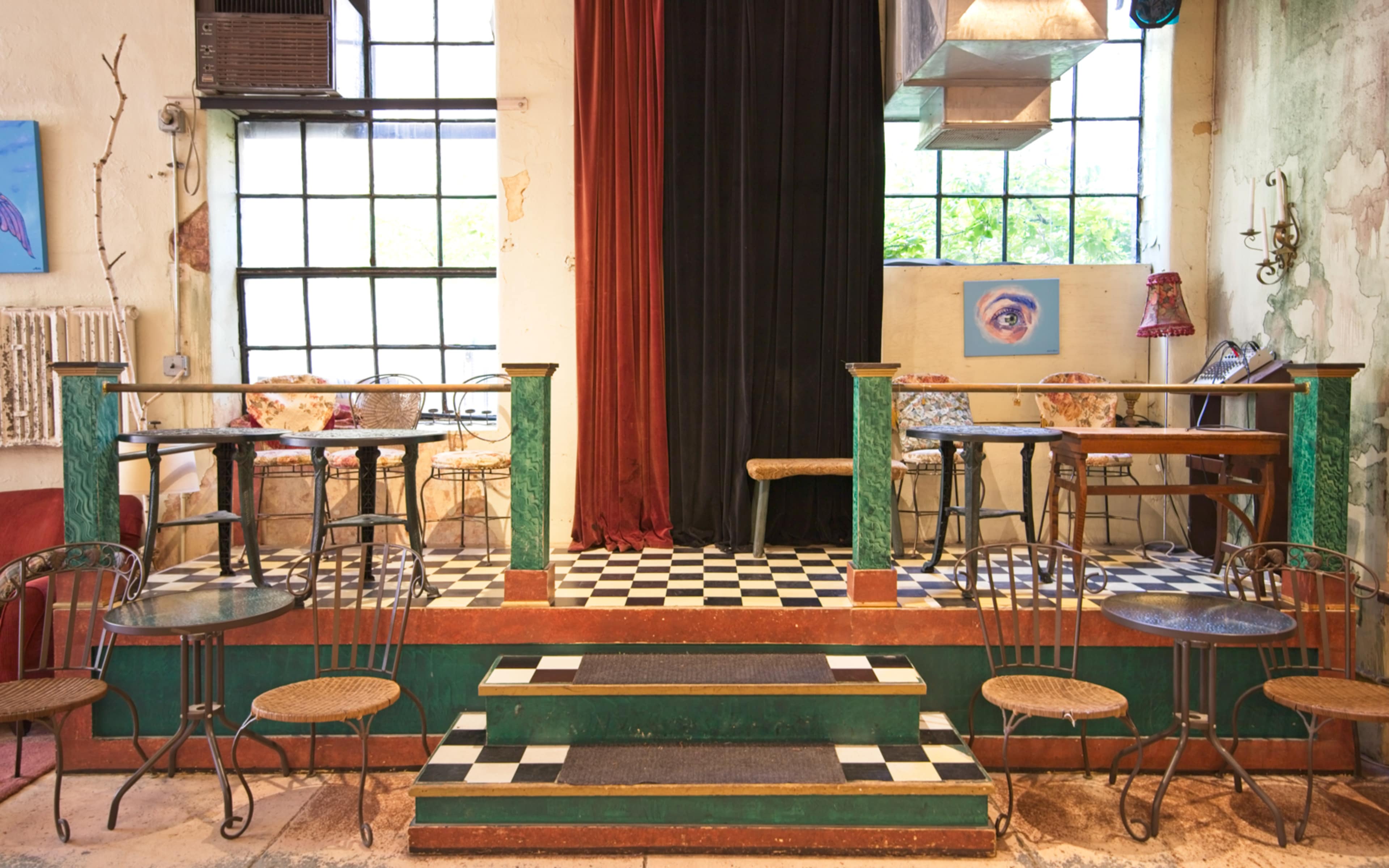An interior of a quaint café featuring a stage area with a checkered floor, surrounded by small tables and chairs against a backdrop of large windows.