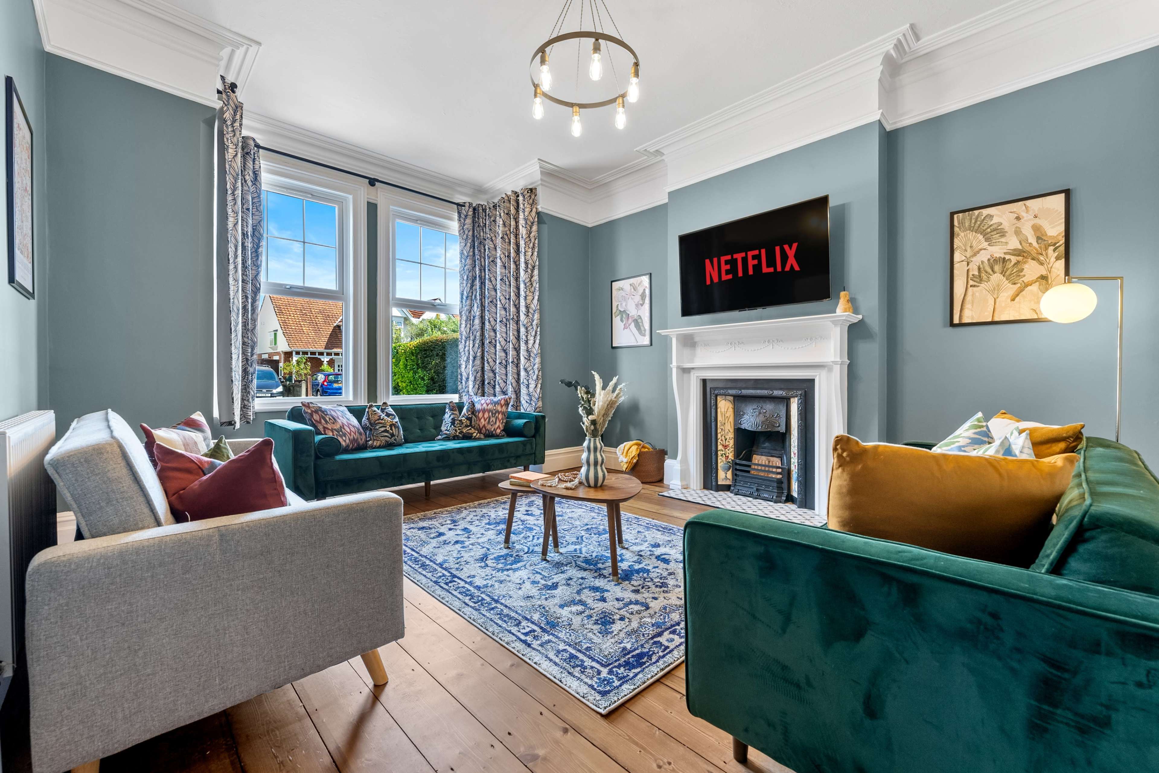A living room features two green velvet sofas and a light gray sofa arranged around a coffee table, with a decorative fireplace and a large screen displaying the Netflix logo on the wall.