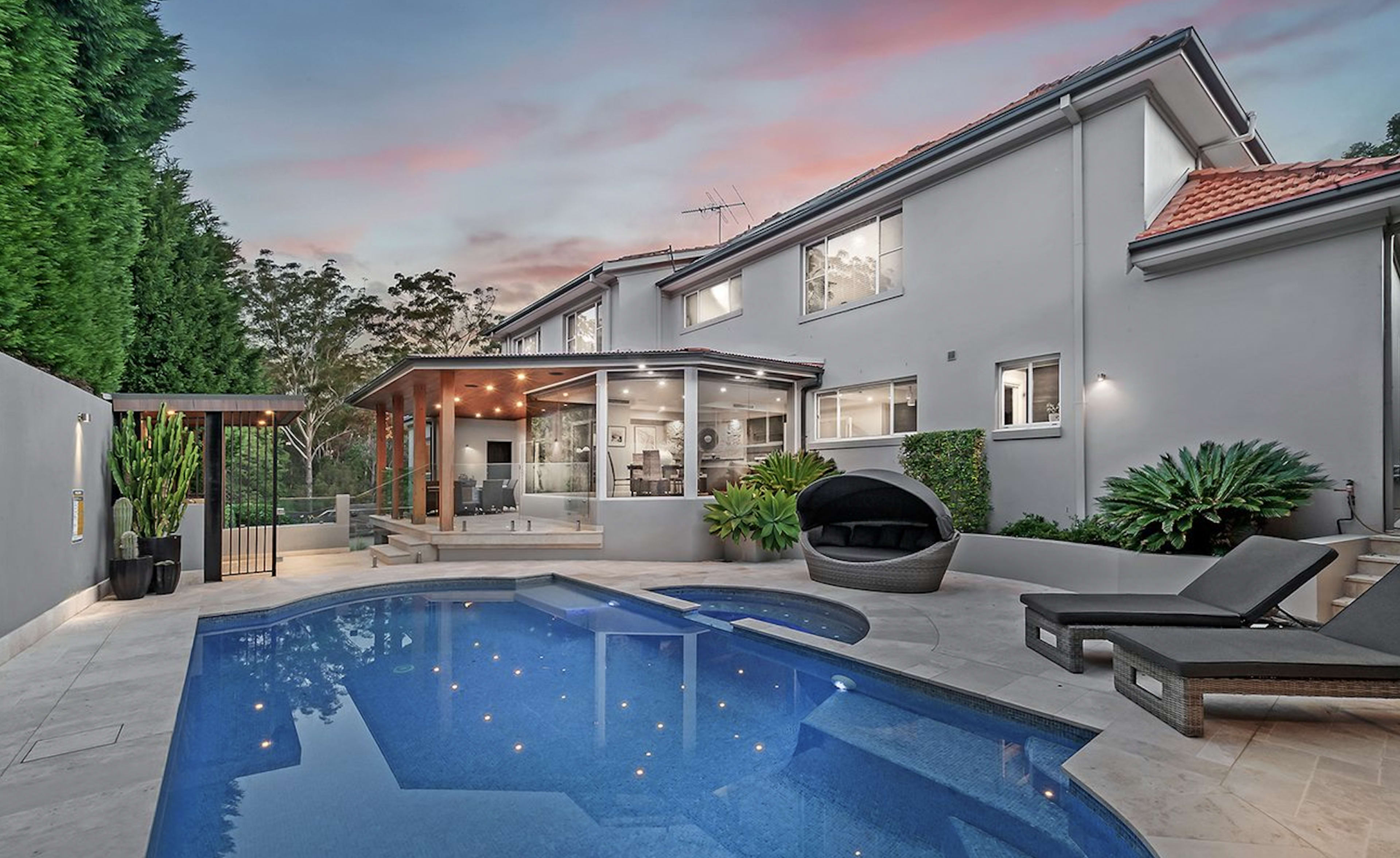 The image shows a modern backyard featuring a swimming pool, lounge chairs, and a covered patio adjacent to a two-story house.