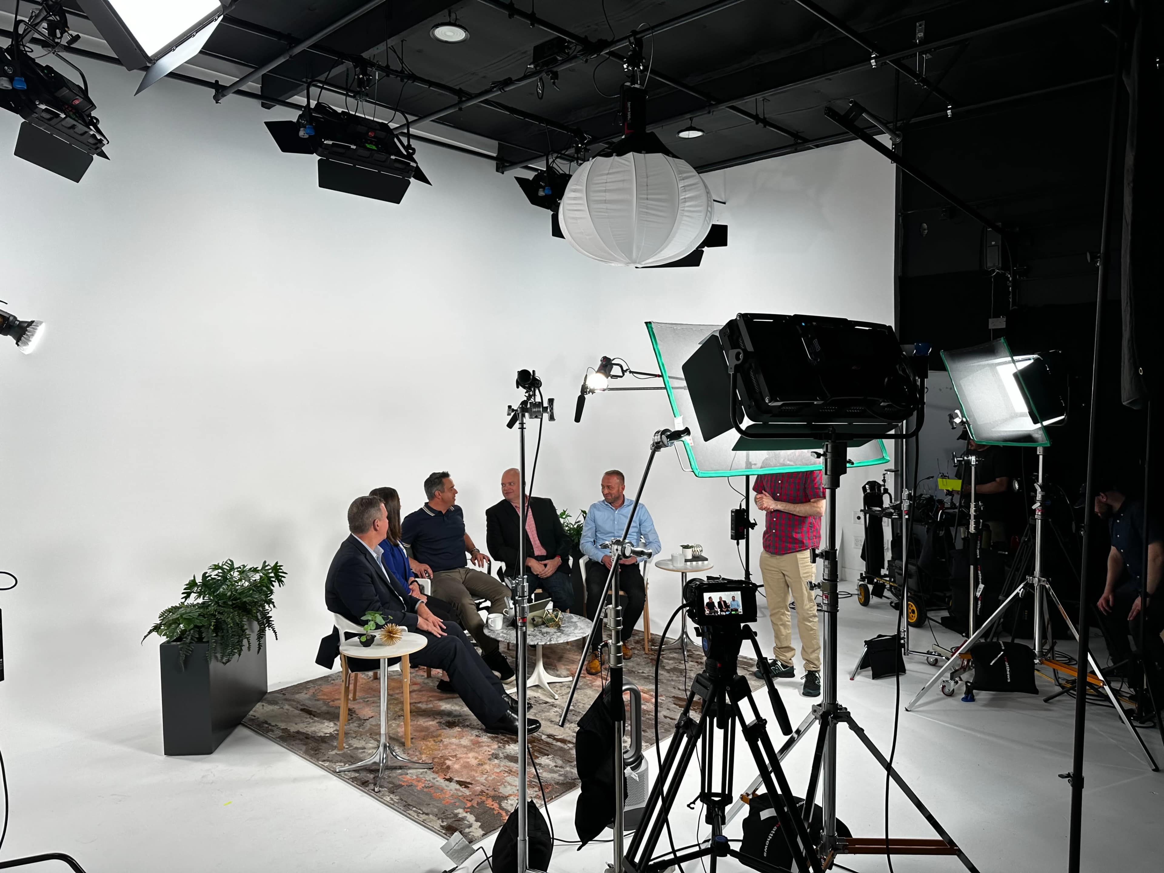 A group of five individuals is seated on a set with a white backdrop, surrounded by various studio lights and camera equipment.