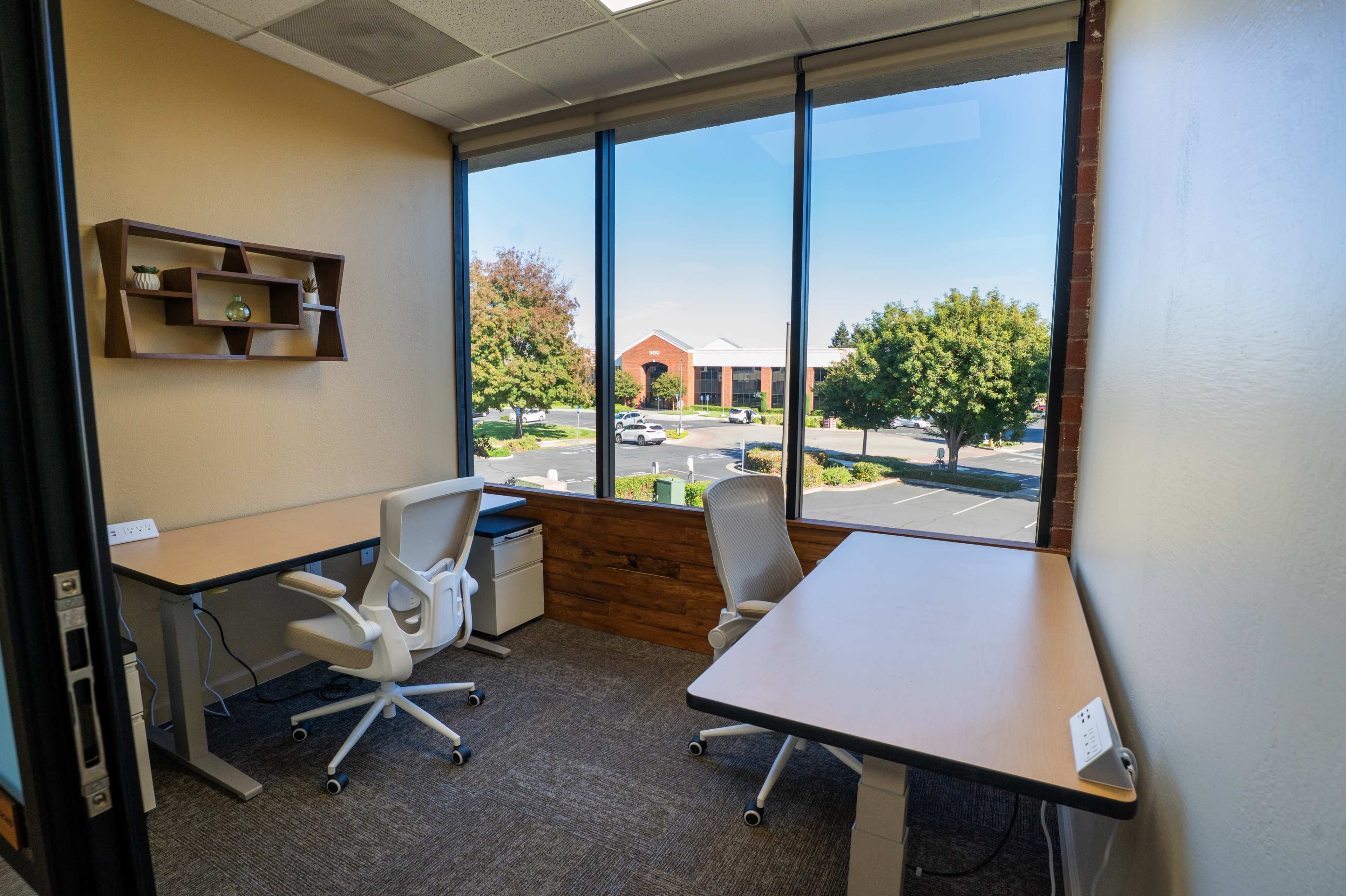 The image shows a small office space with two desks and ergonomic chairs, featuring large windows that overlook a parking lot and landscaped area outside.