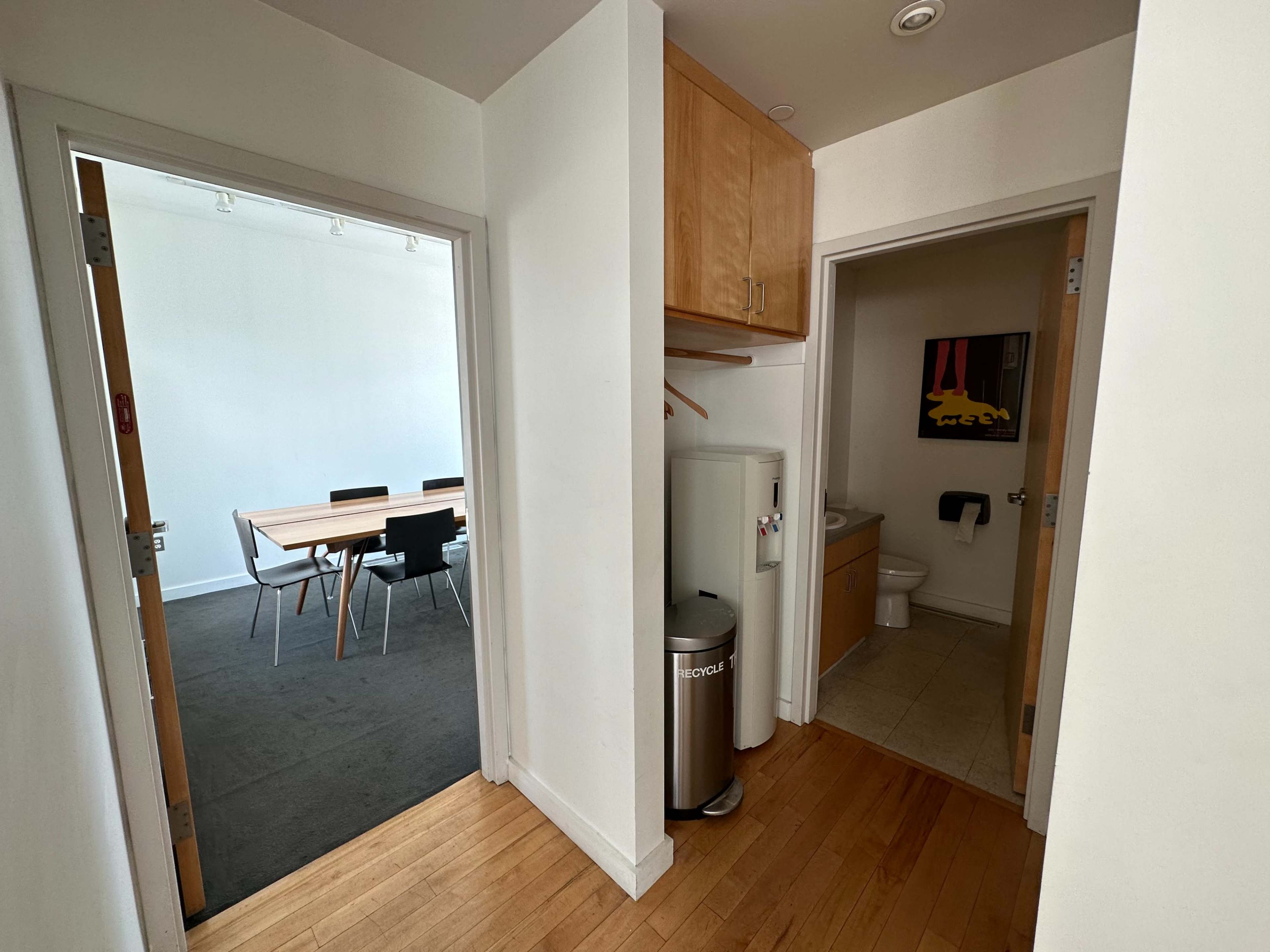 A hallway with two doorways leading to a meeting room on the left and a bathroom on the right, featuring a refrigerator and a trash can near the bathroom entrance.