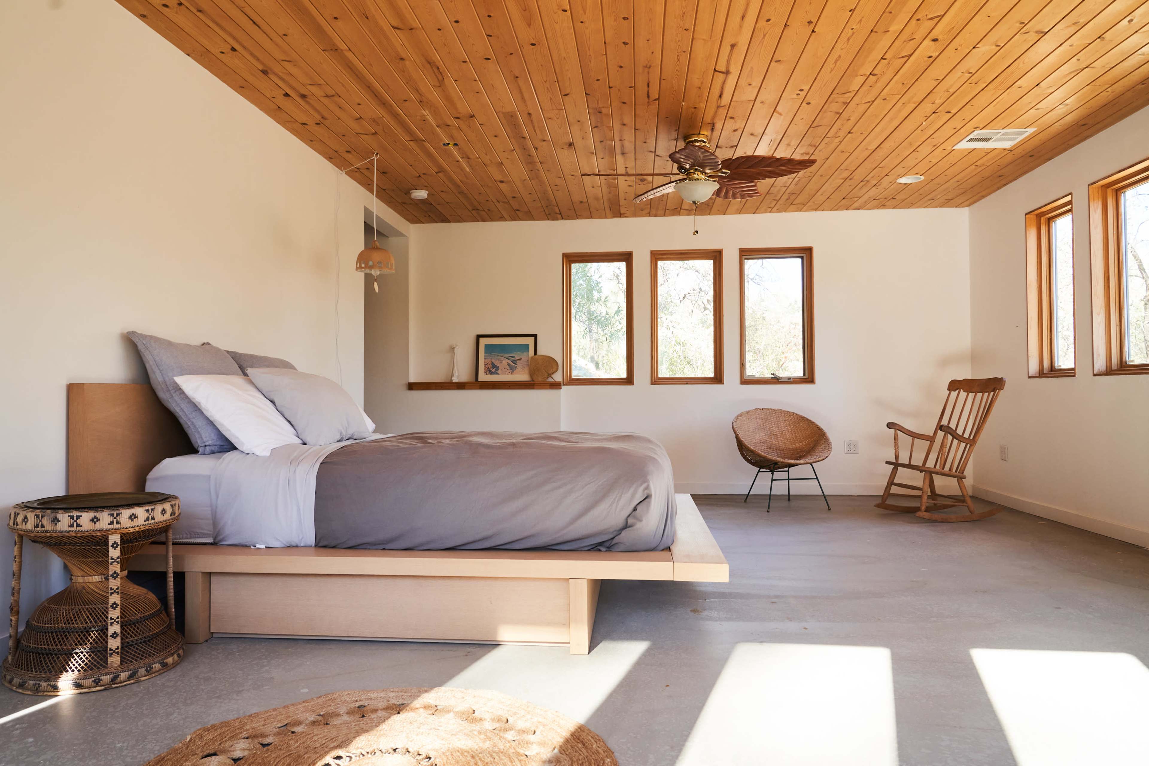 A minimalist bedroom features a wooden ceiling, a bed with gray bedding, a rocking chair, and three windows allowing natural light.