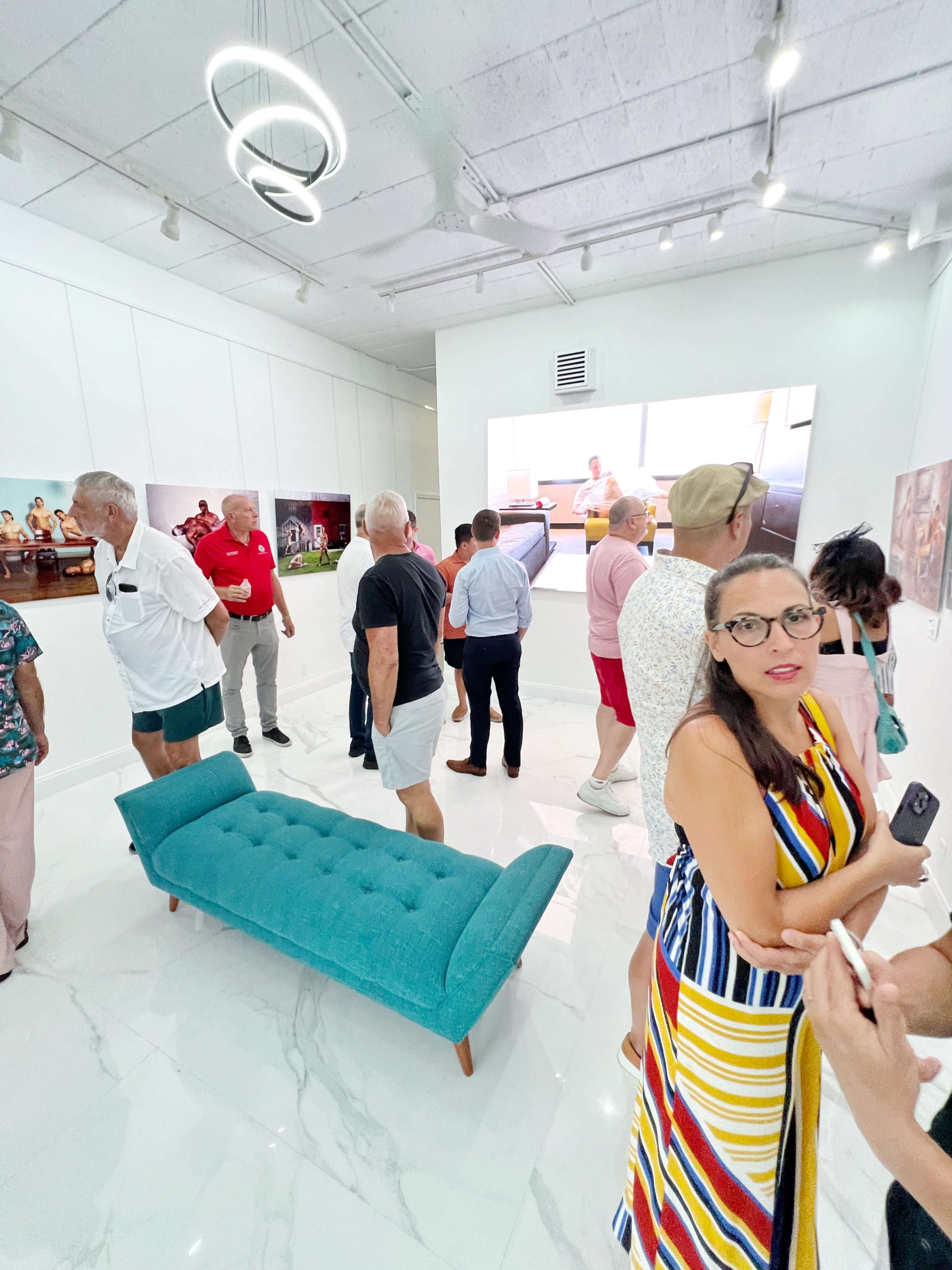 A group of people is gathered in a modern gallery, with a large screen displaying an image and a teal bench in the center of the room.
