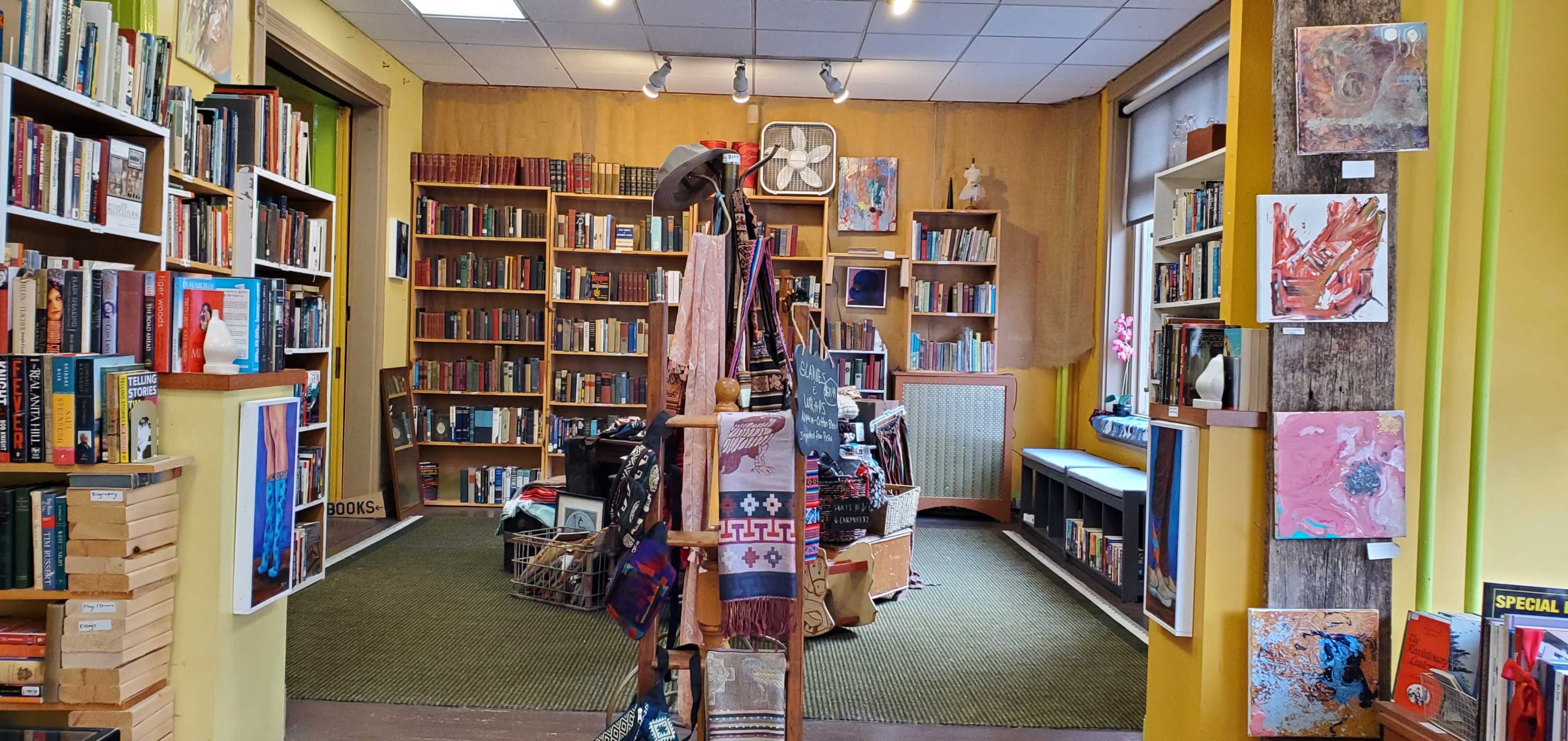 An interior of a bookstore with bookshelves filled with books, artwork on the walls, and a display of scarves and items in the foreground.
