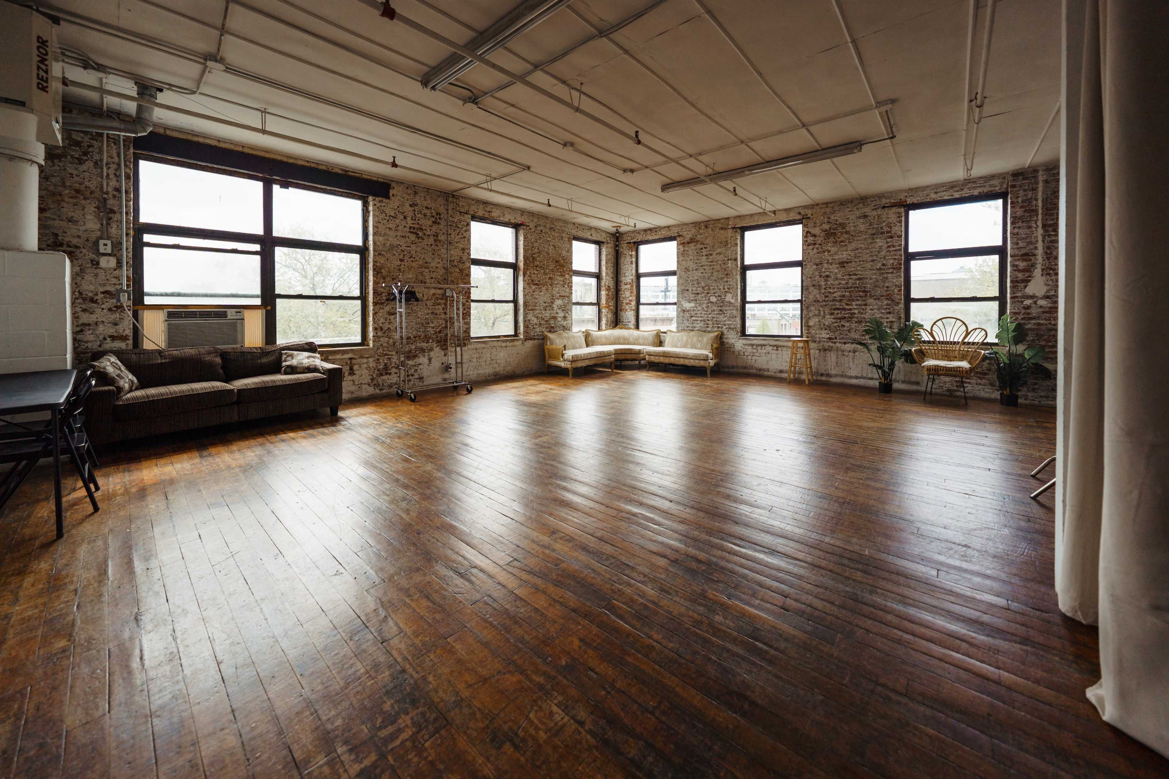A spacious, well-lit room with large windows, wooden floors, and minimal furnishings, including a couch and a chair.