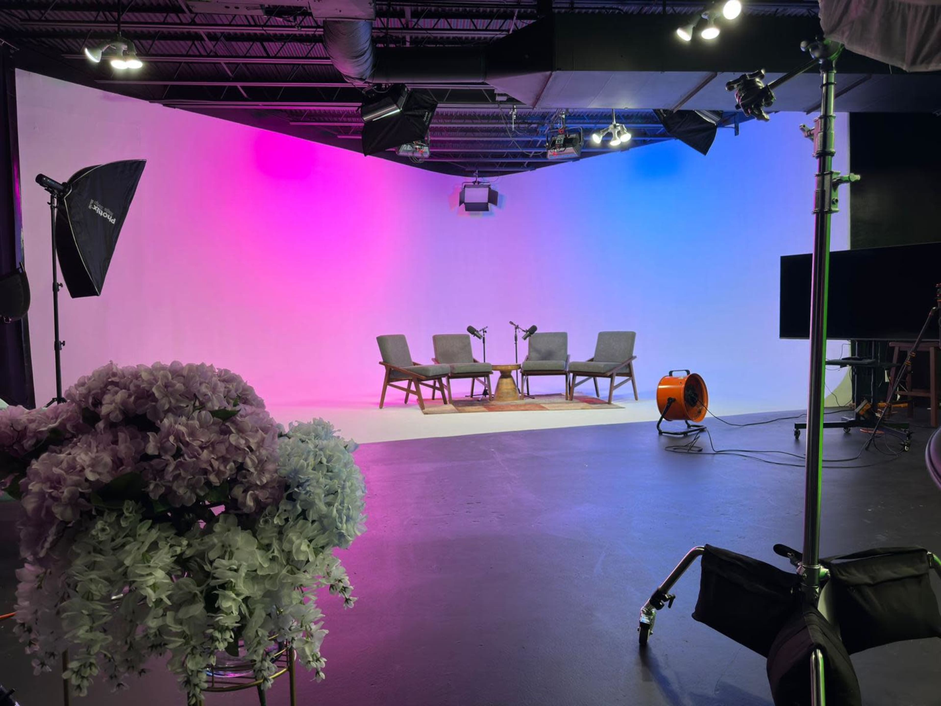 A photography studio features a brightly lit set with three chairs, a small table, and a backdrop that transitions from pink to blue.