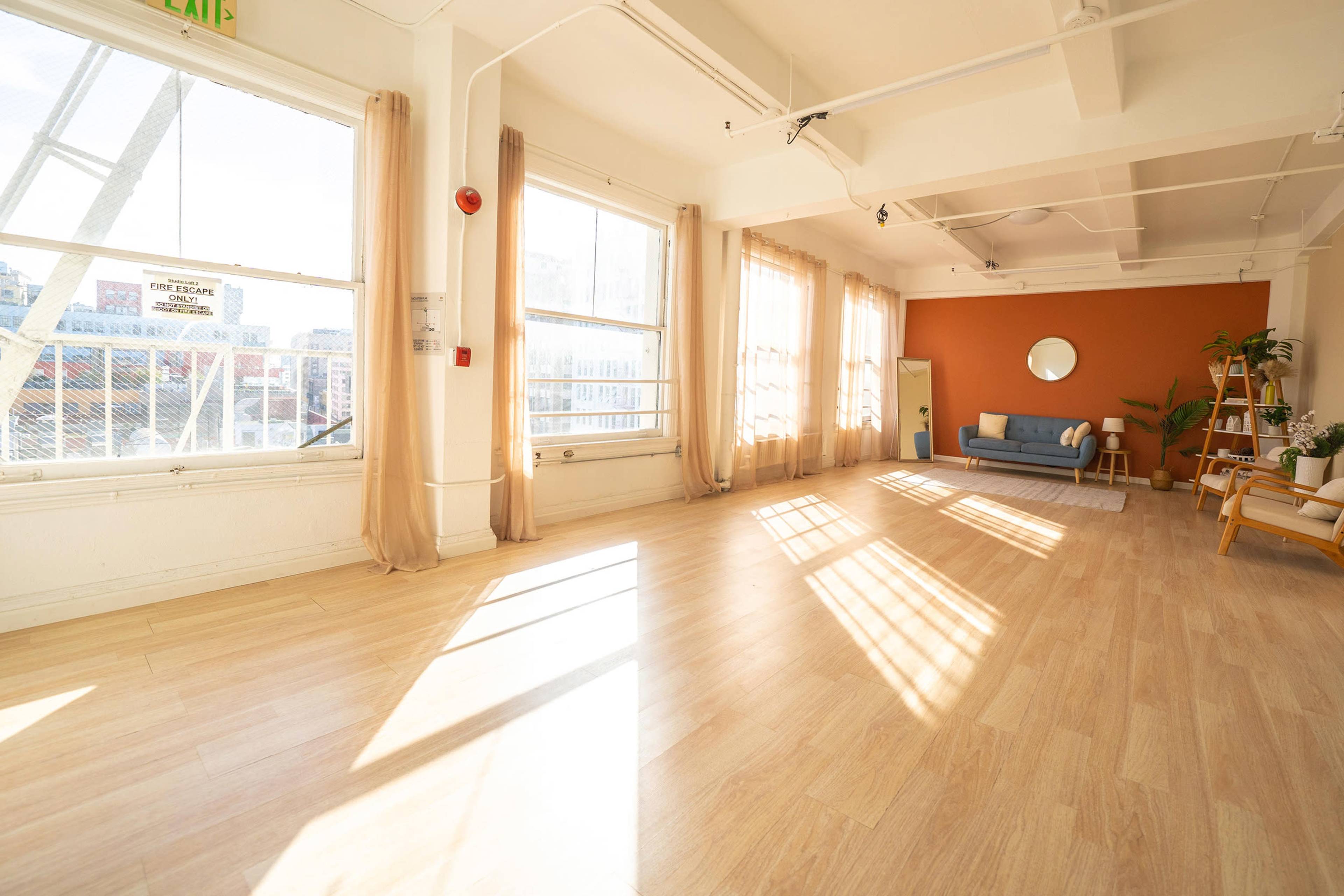 A bright, empty studio space with large windows, light wood flooring, and minimal furniture, including a sofa and a chair against a wall with an orange accent.