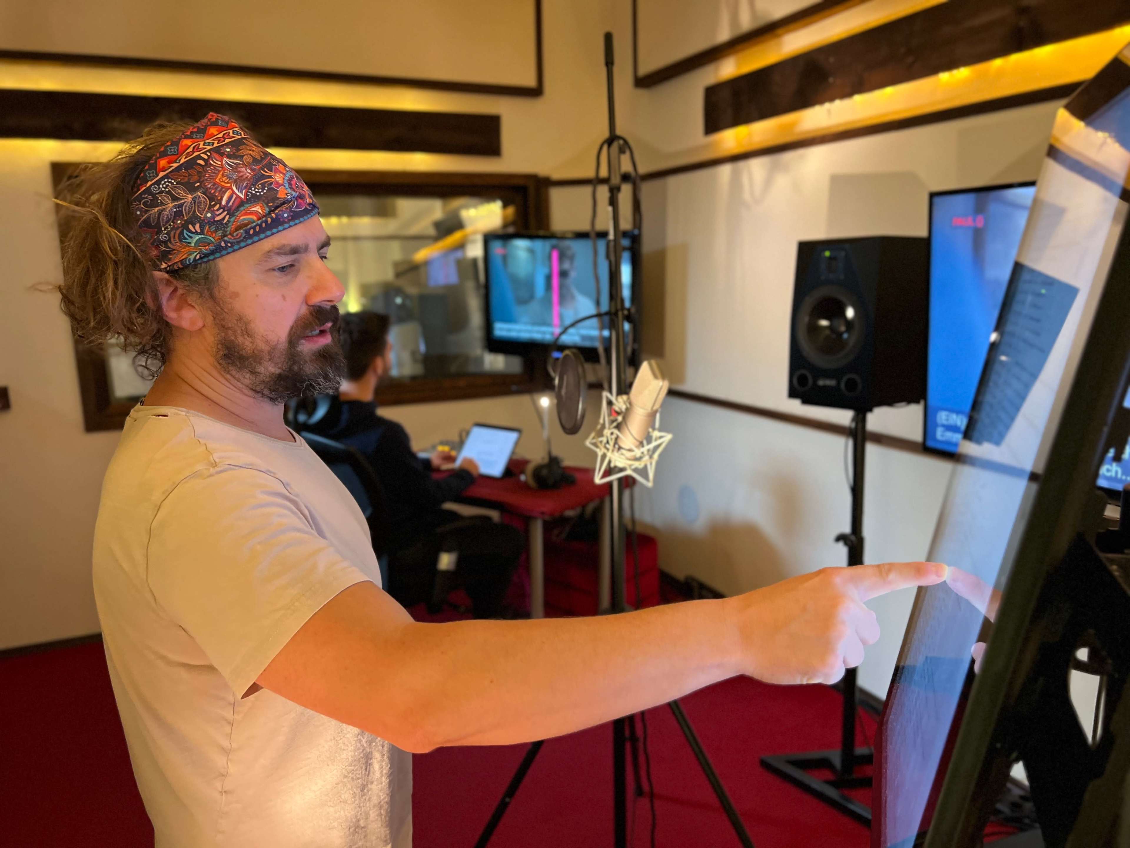 A man with curly hair and a bandana is pointing at a touchscreen while standing in a recording studio, with another person working on a laptop in the background.
