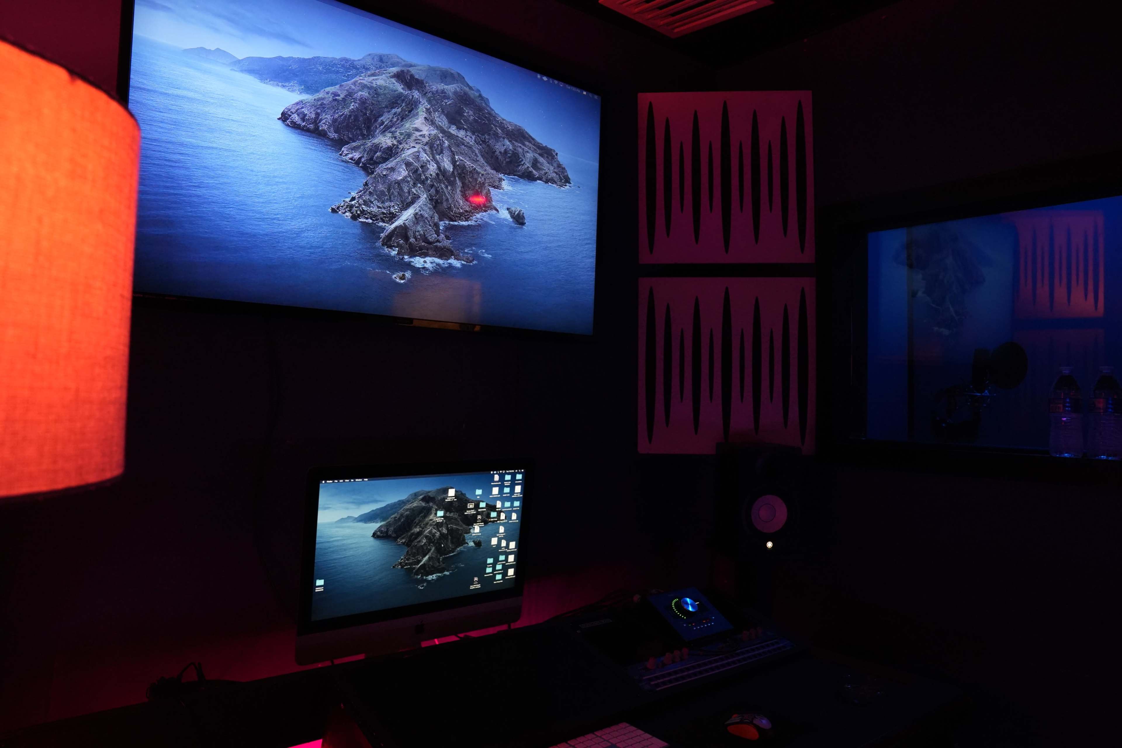 A dimly lit recording studio features a large screen displaying a coastal landscape, a computer monitor with a similar image, and soundproofing panels on the walls.