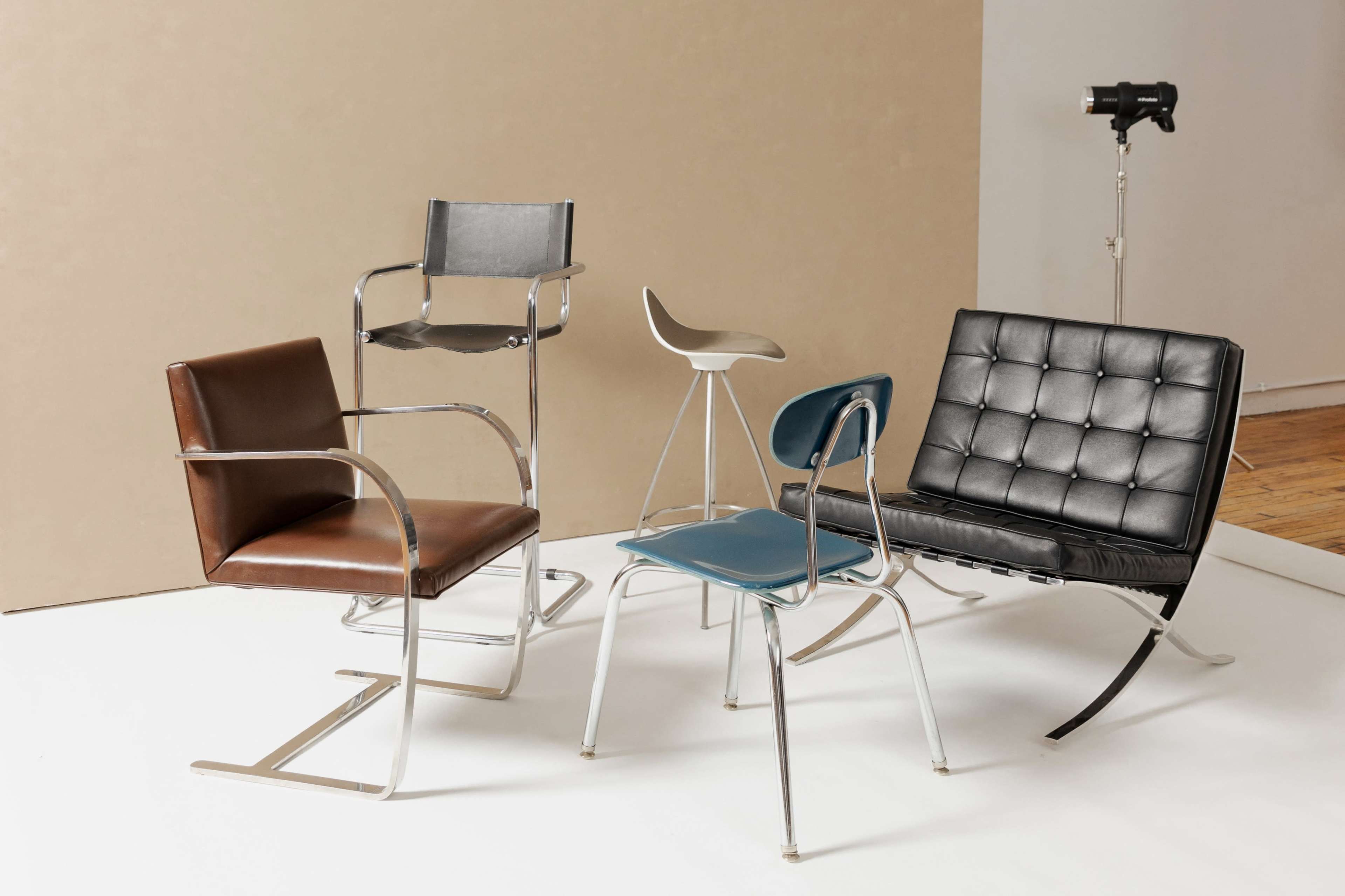 The image shows a collection of four distinct chairs arranged on a white floor, with a tan background and a camera on a tripod nearby.