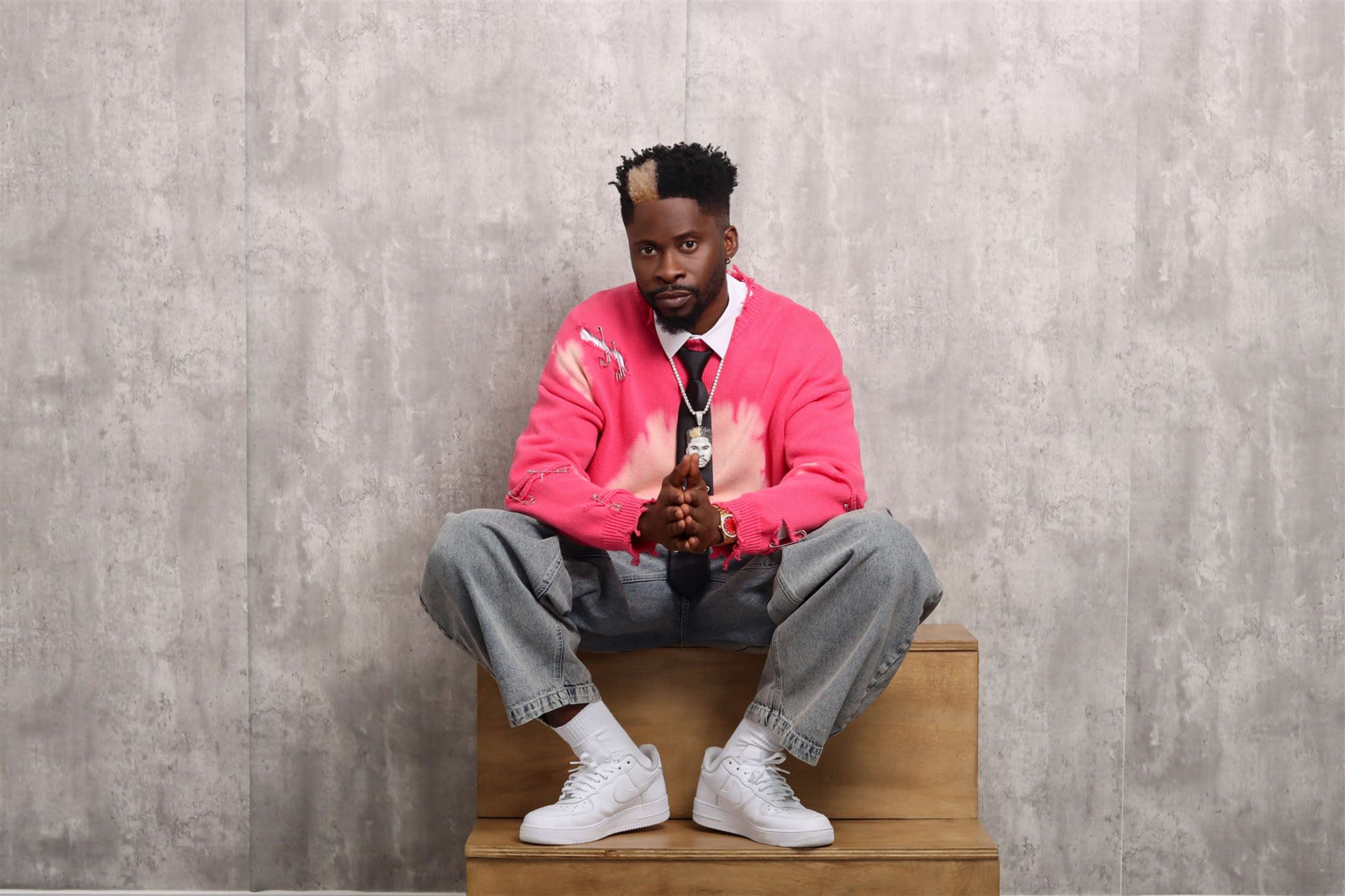 A person sits on a wooden platform, wearing a pink tie-dye sweater, gray jeans, and white sneakers, with their hands clasped and a neutral expression.