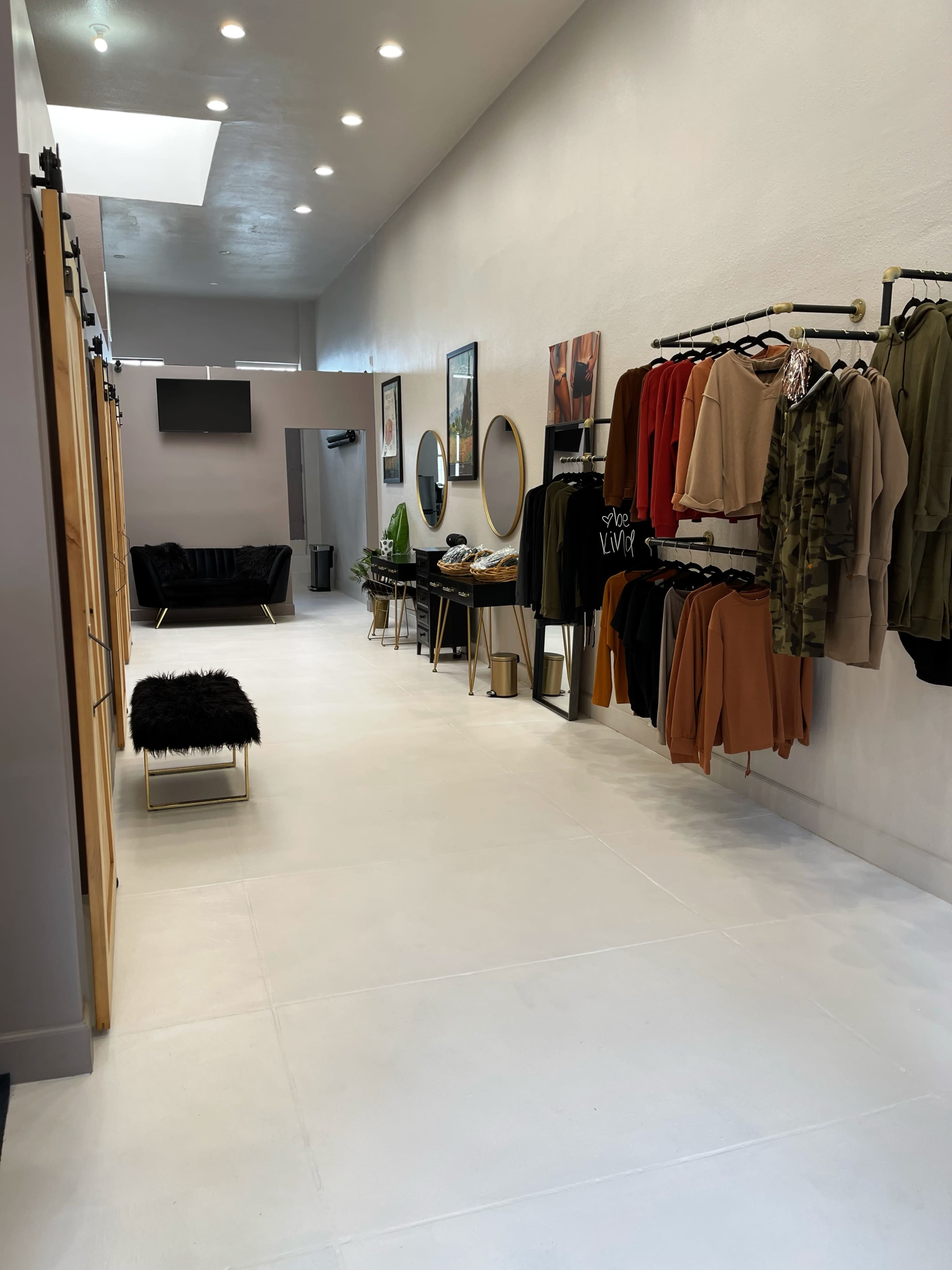 The image shows a modern retail space with clothing racks displaying various garments, a seating area, and large mirrors along a well-lit white floor.
