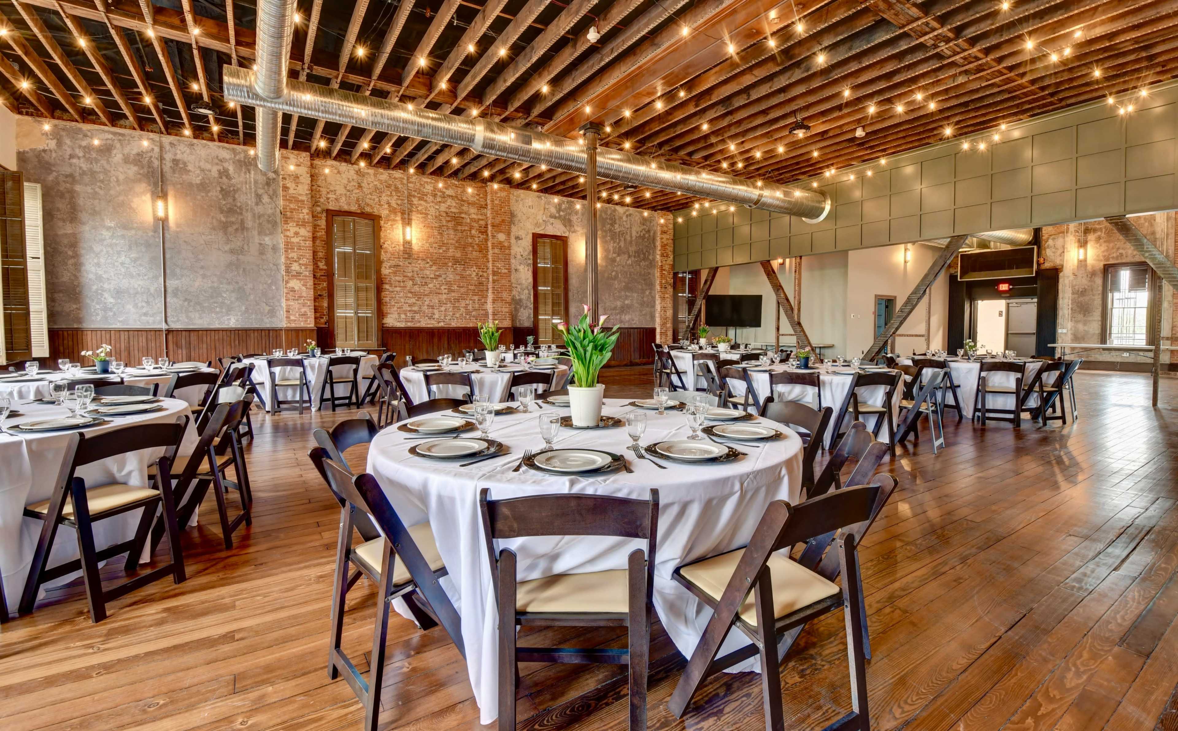 A spacious dining area with round tables set with white tablecloths, black chairs, and decorative centerpieces, under exposed wooden beams and string lights.