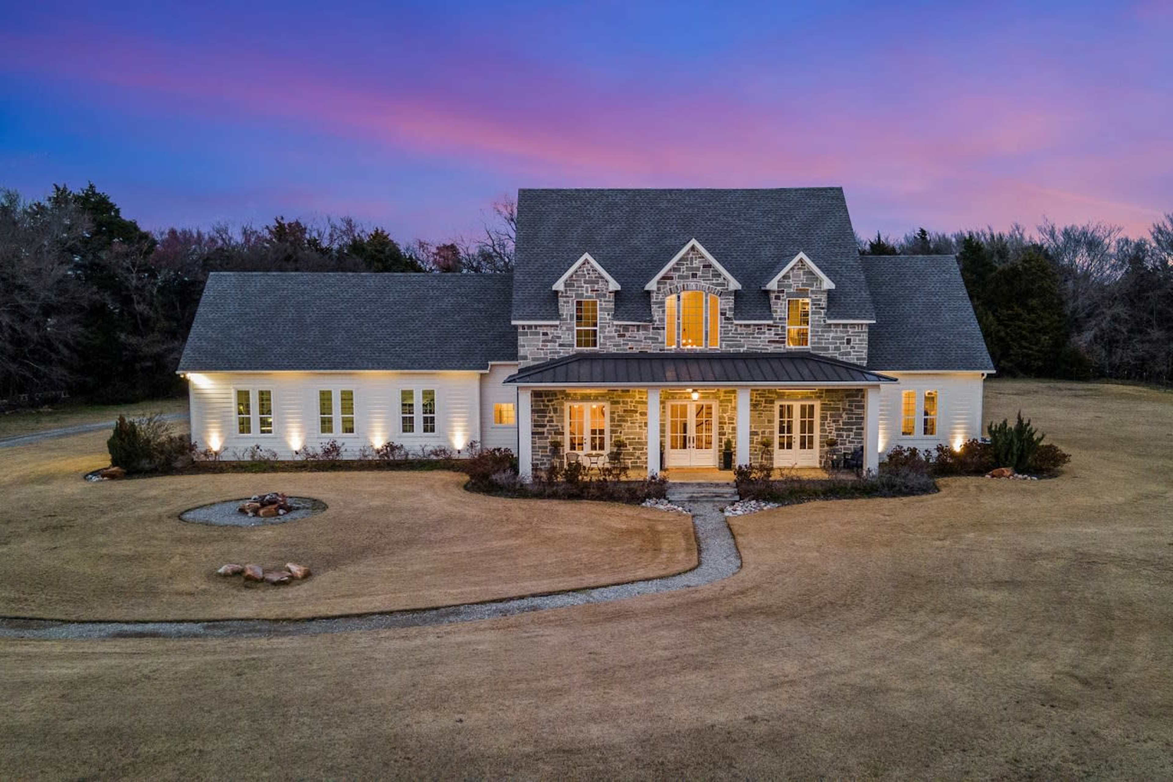 A large stone and wood house sits on a grassy lot, illuminated by exterior lights under a colorful sunset sky.