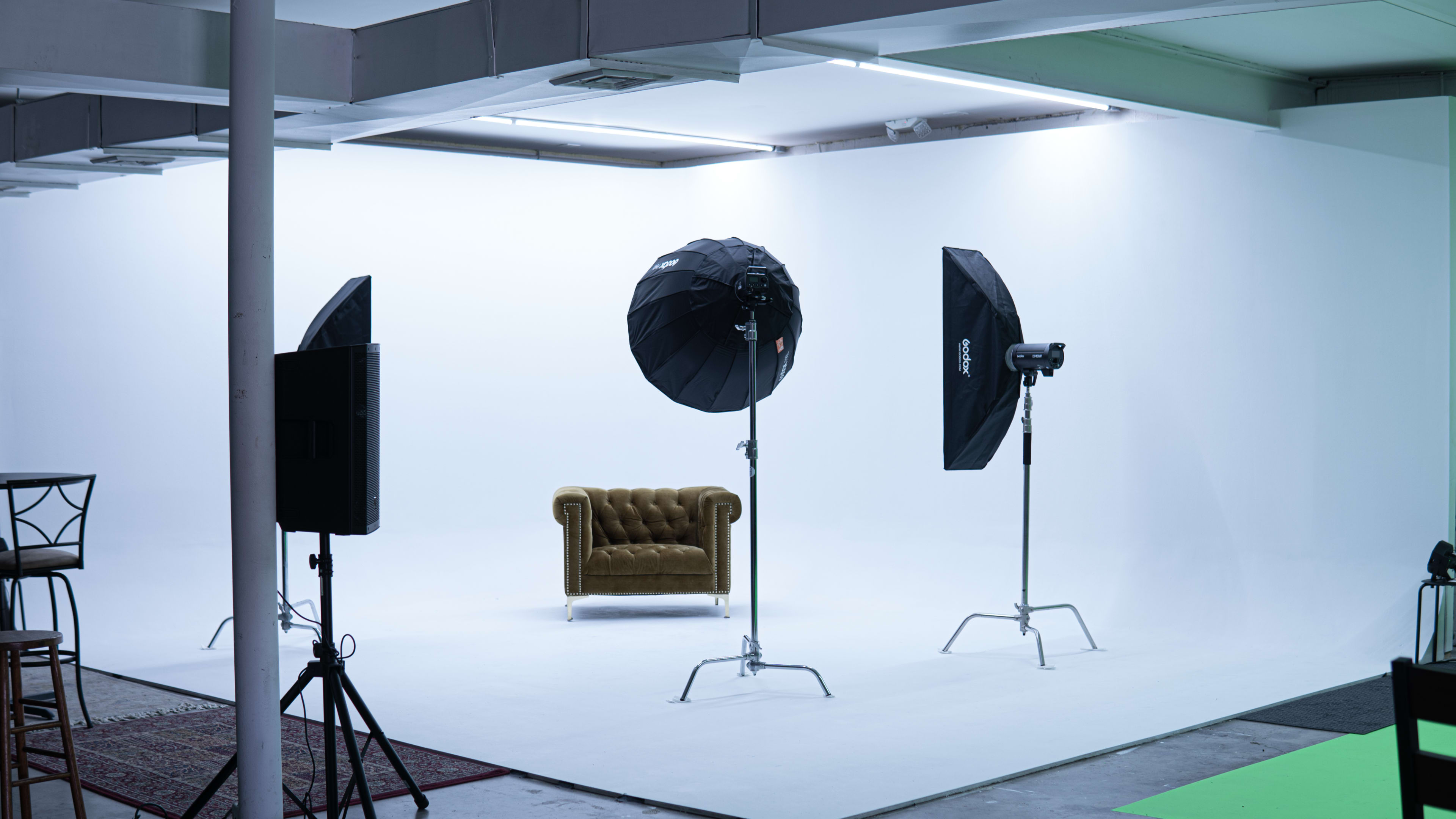 A photography studio features a white backdrop, a brown tufted armchair, and two large softbox lights on stands.