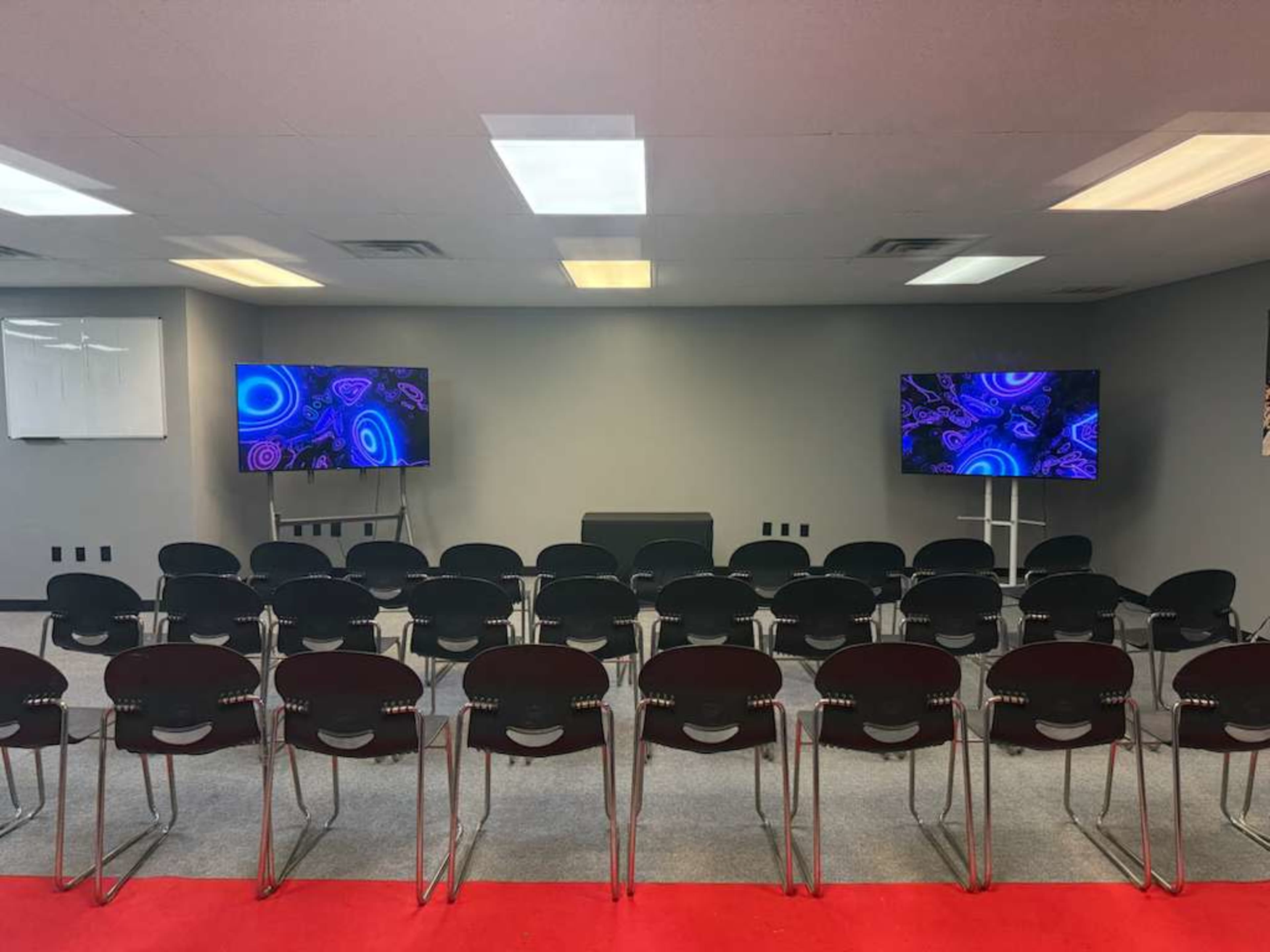 The image shows a room set up for a presentation, featuring two screens on the walls and rows of black chairs facing the screens.