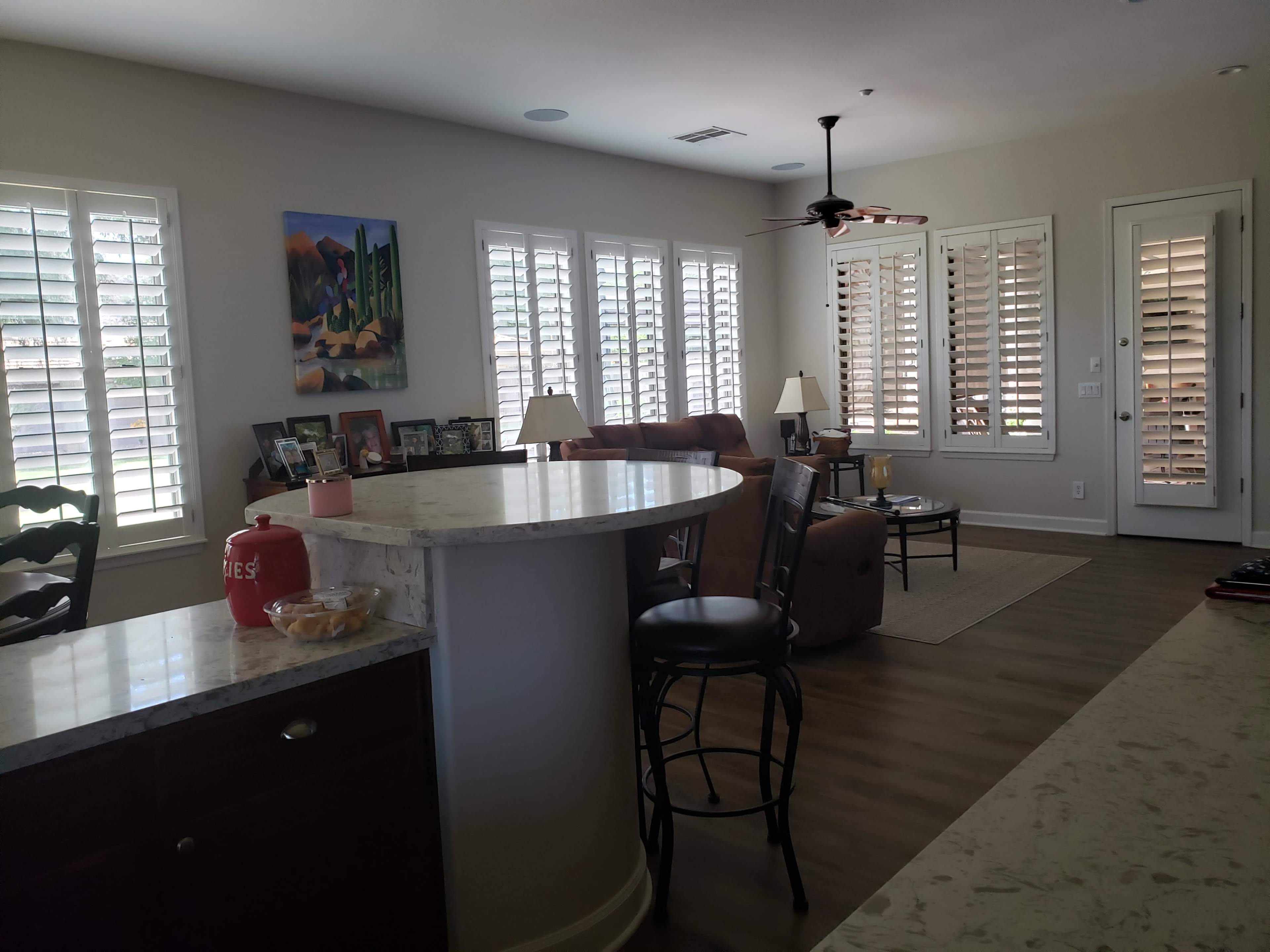 A living room connected to a kitchen area with a bar stool setup, featuring large windows with shutters and light-colored walls.