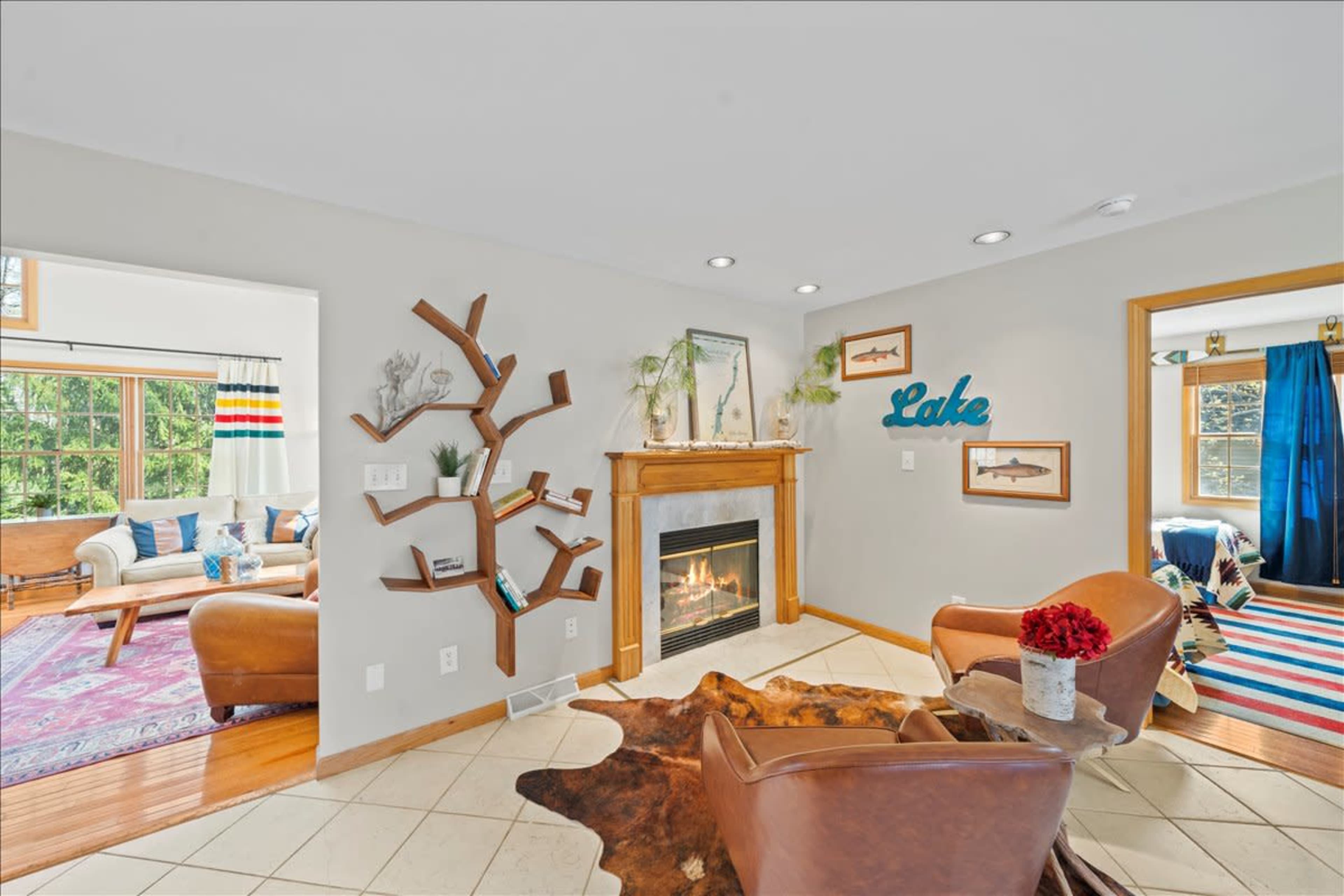 A cozy living room featuring a fireplace, wooden furnishings, and decorative wall accents, with views of a lake visible through a nearby window.