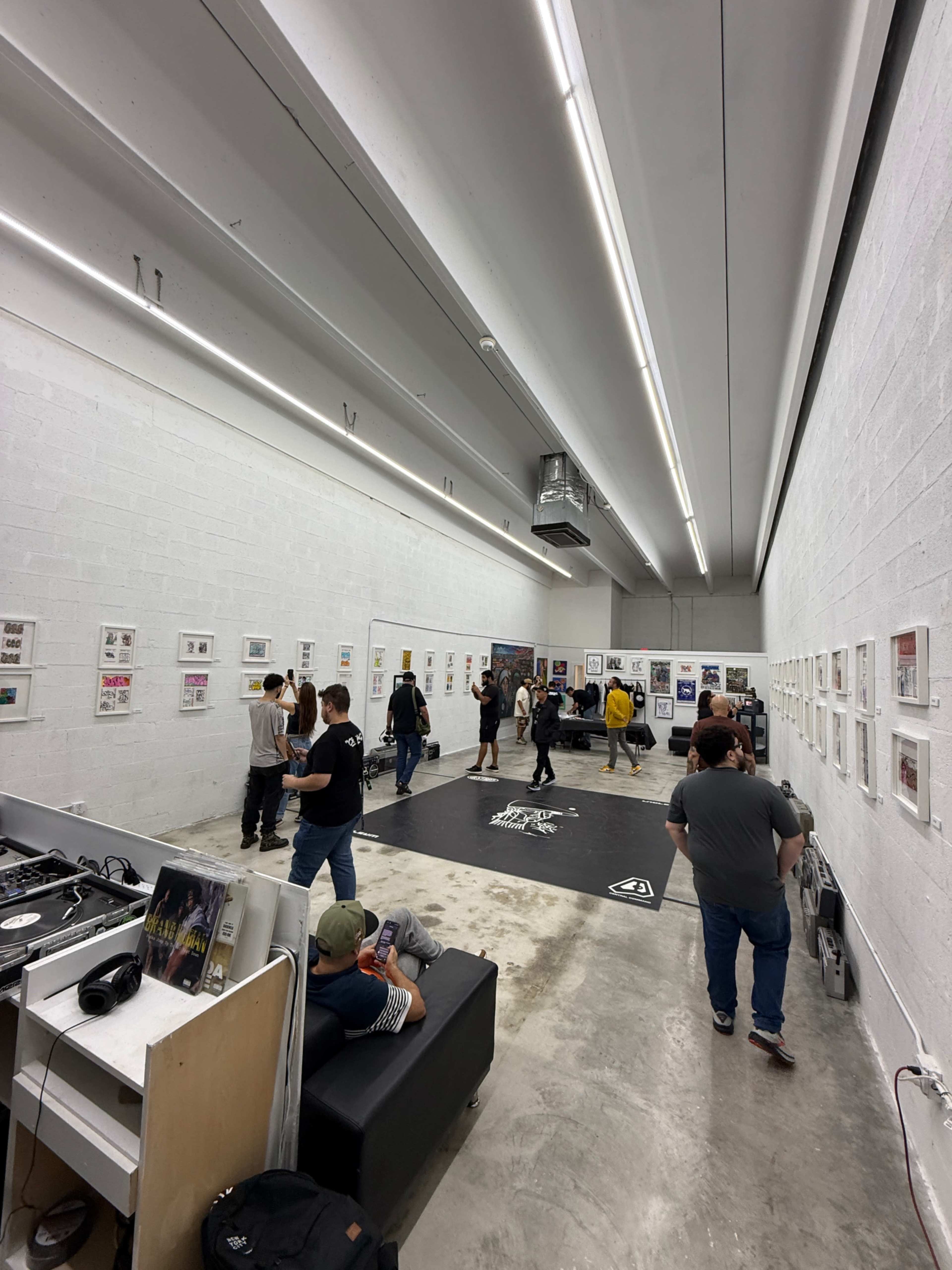 The image shows a spacious art gallery filled with people engaging with framed artworks on the walls, while a DJ setup is visible in the corner.