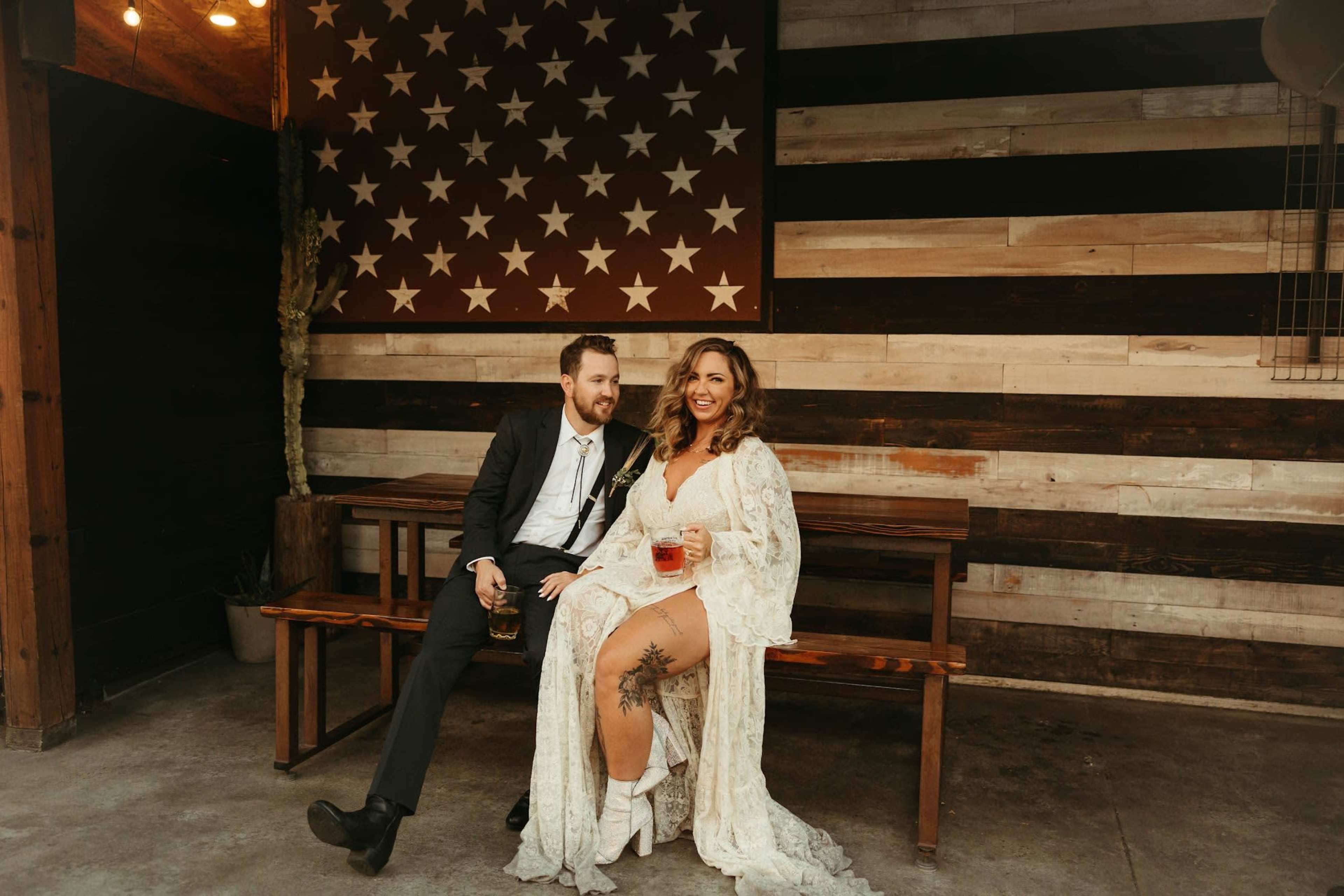 A couple sits on a wooden bench, holding drinks, in front of a large American flag painted on a wooden wall.