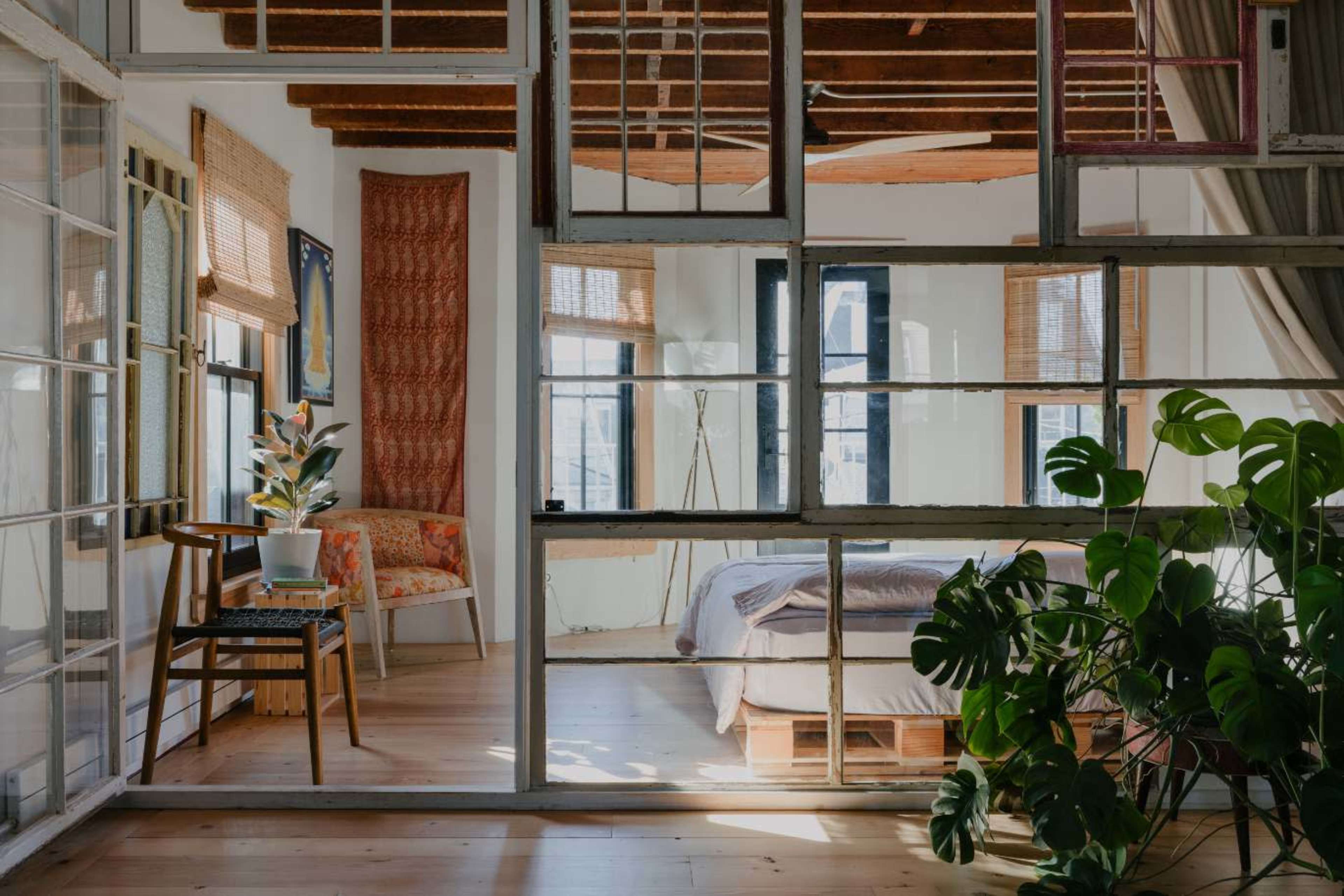 A bright, airy bedroom with large windows, a plant in the foreground, and a cozy seating area visible through a glass partition.