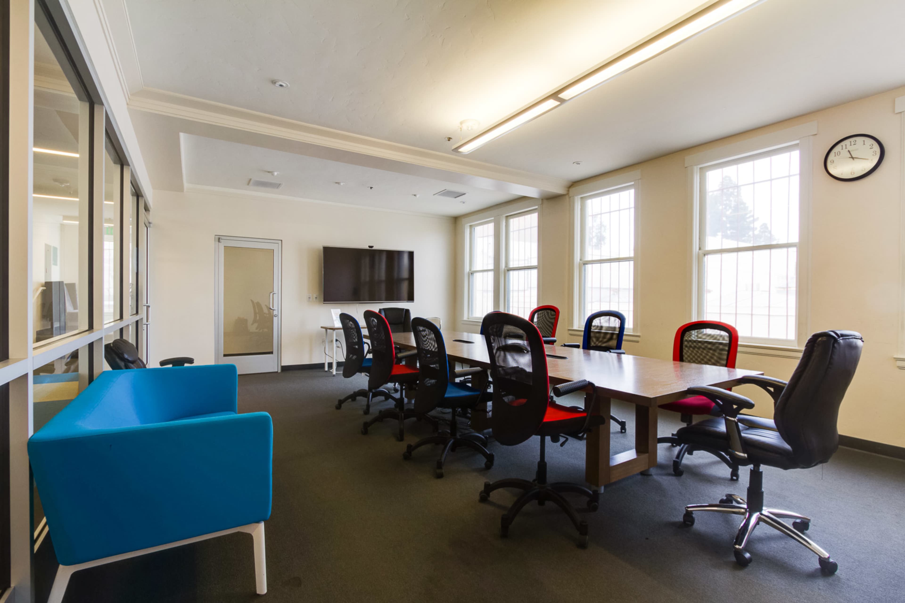 A conference room features a long table surrounded by several office chairs, a television on the wall, and a blue sofa in the corner.