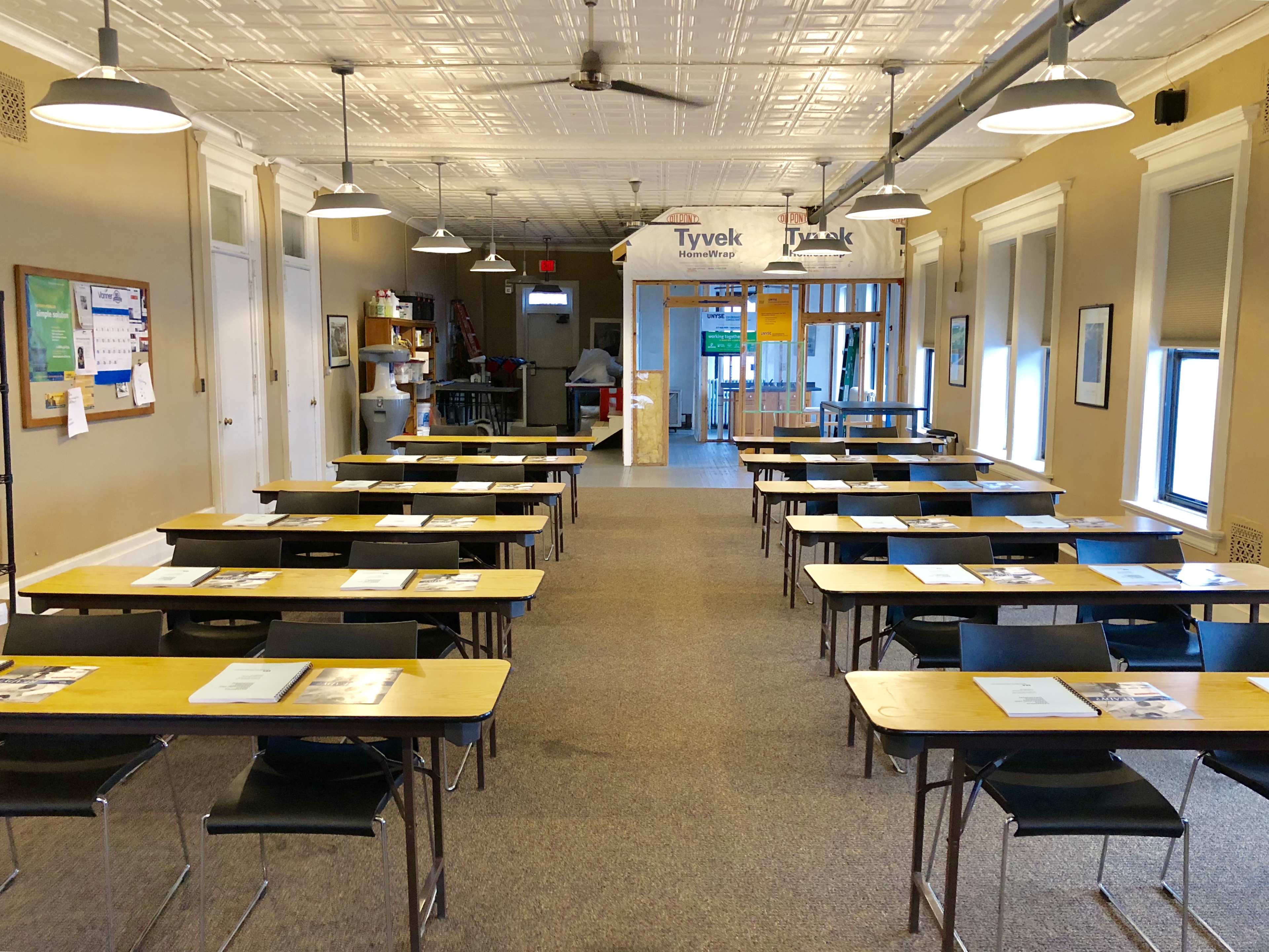 A classroom arranged with several rows of tables and chairs facing the front, with a bulletin board and a small kitchenette visible in the background.