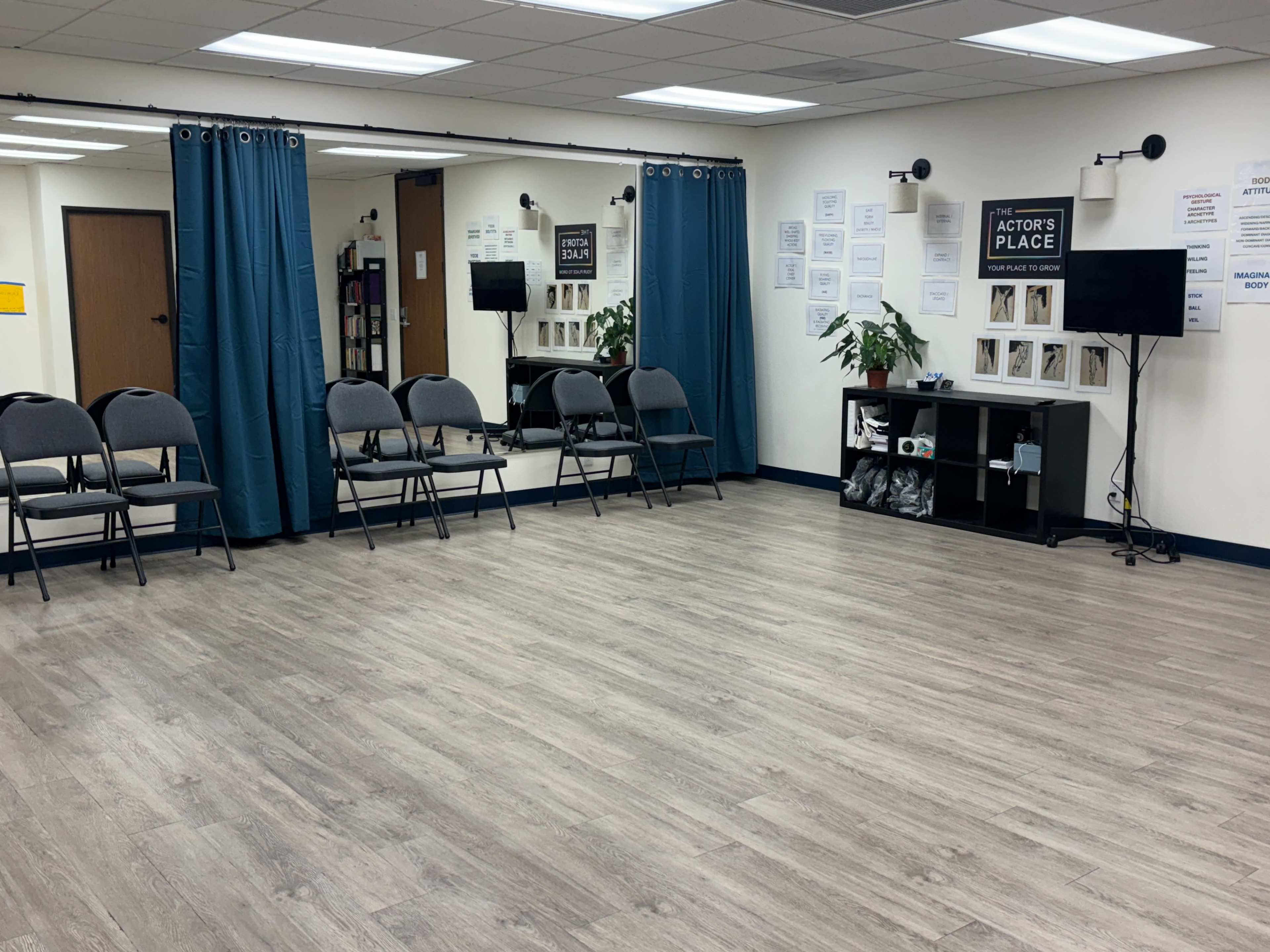 The image shows a spacious rehearsal room with blue curtains, several arranged chairs, a mirrored wall, and a black shelving unit displaying framed photographs.