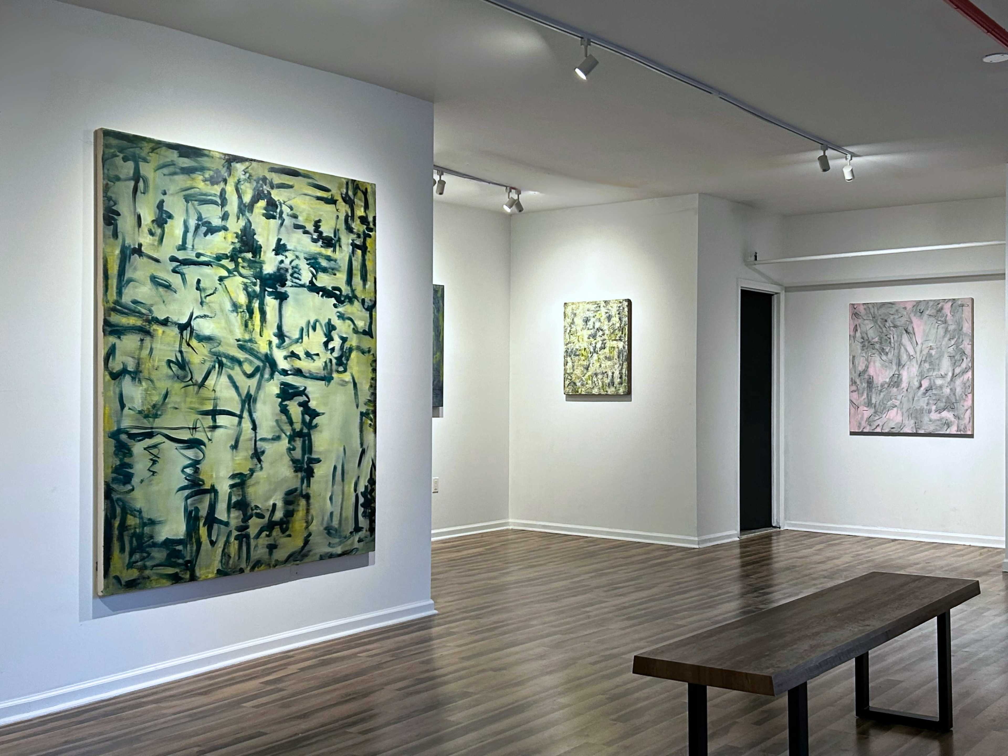 The image shows an art gallery interior featuring several abstract paintings on the walls and a wooden bench in the center of the space.
