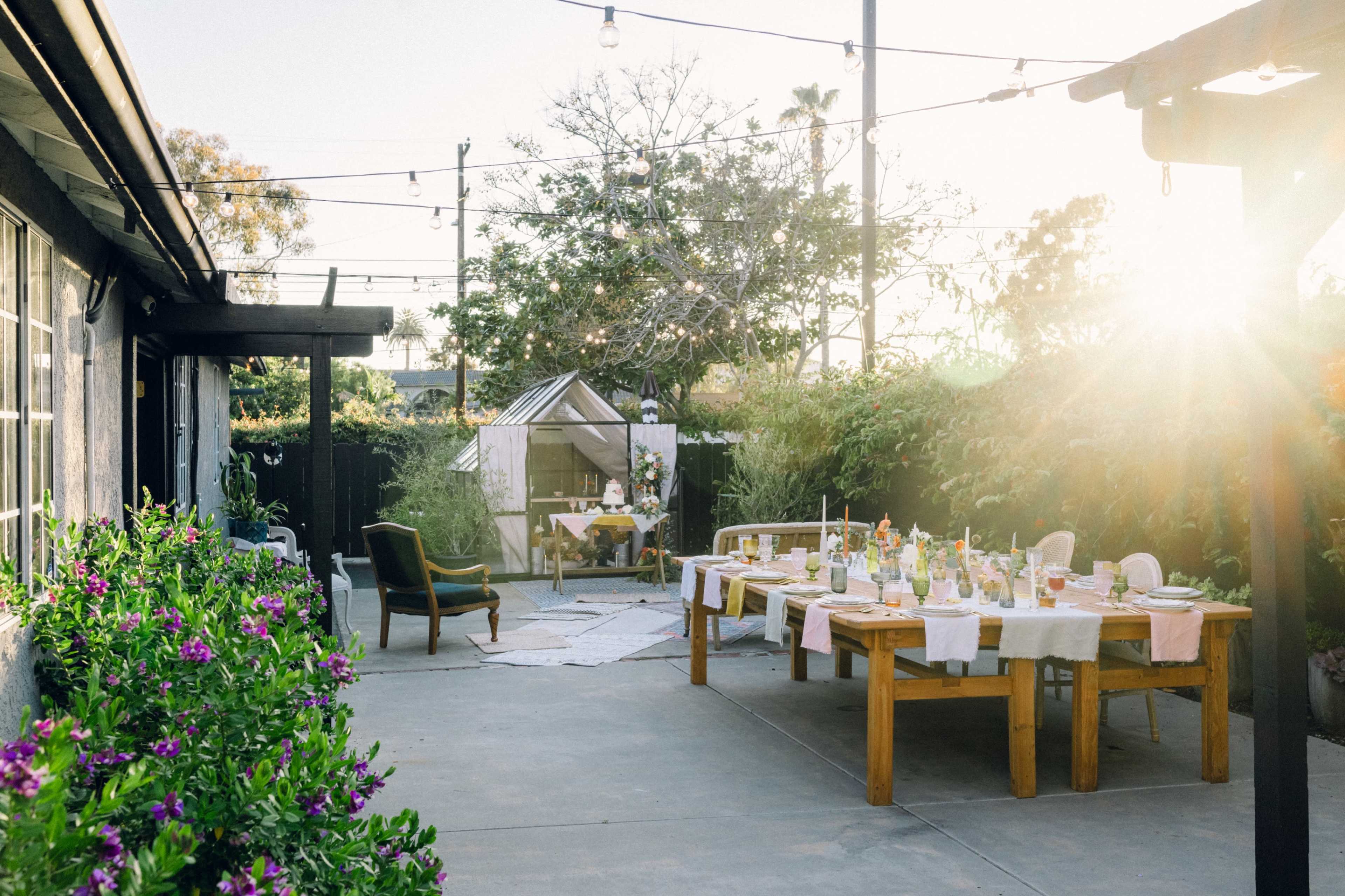 A long dining table is set outdoors in a garden with string lights overhead and a greenhouse in the background.
