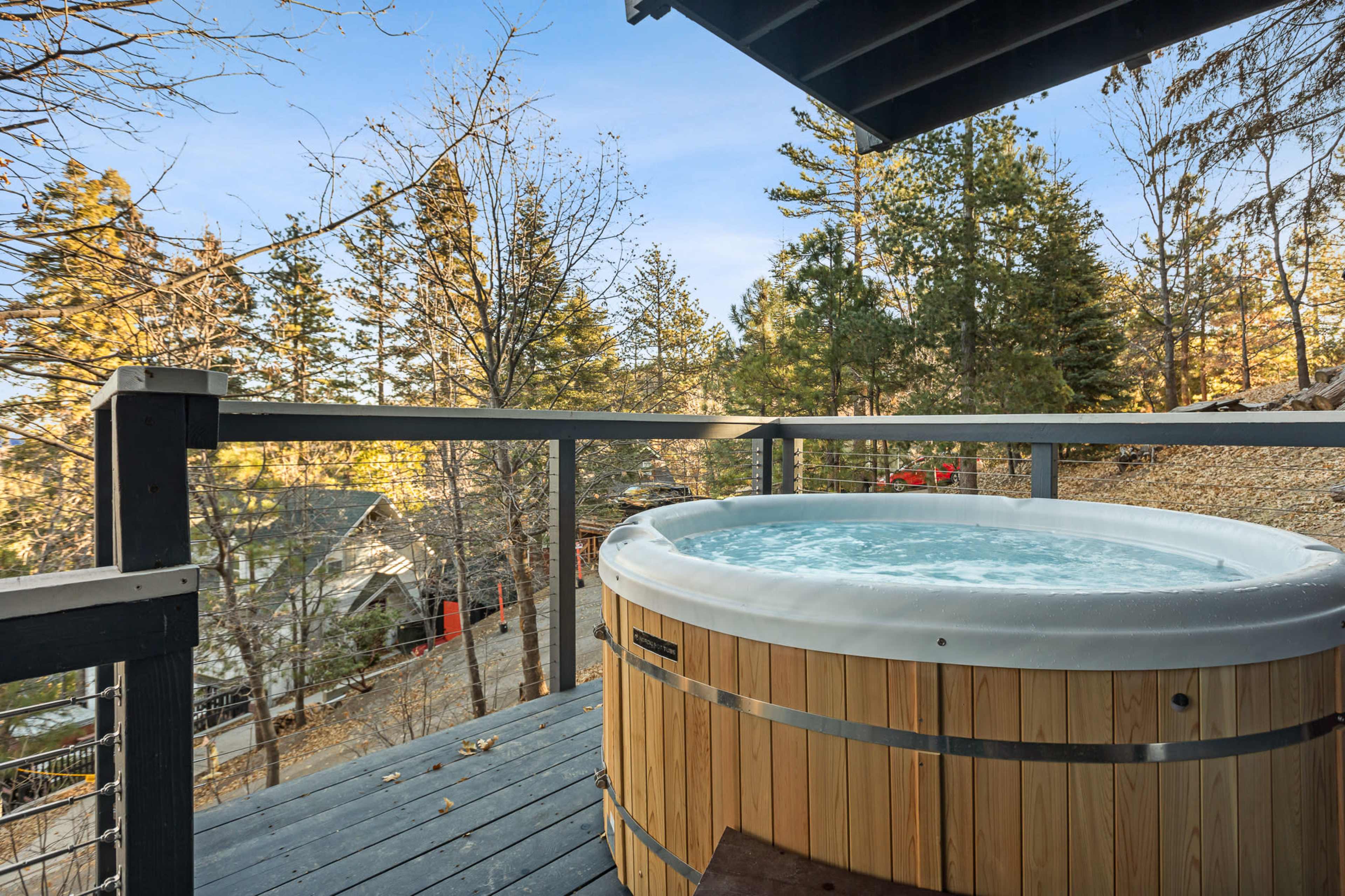 A hot tub made of wood is situated on a balcony surrounded by trees and overlooking residential buildings in a tranquil setting.