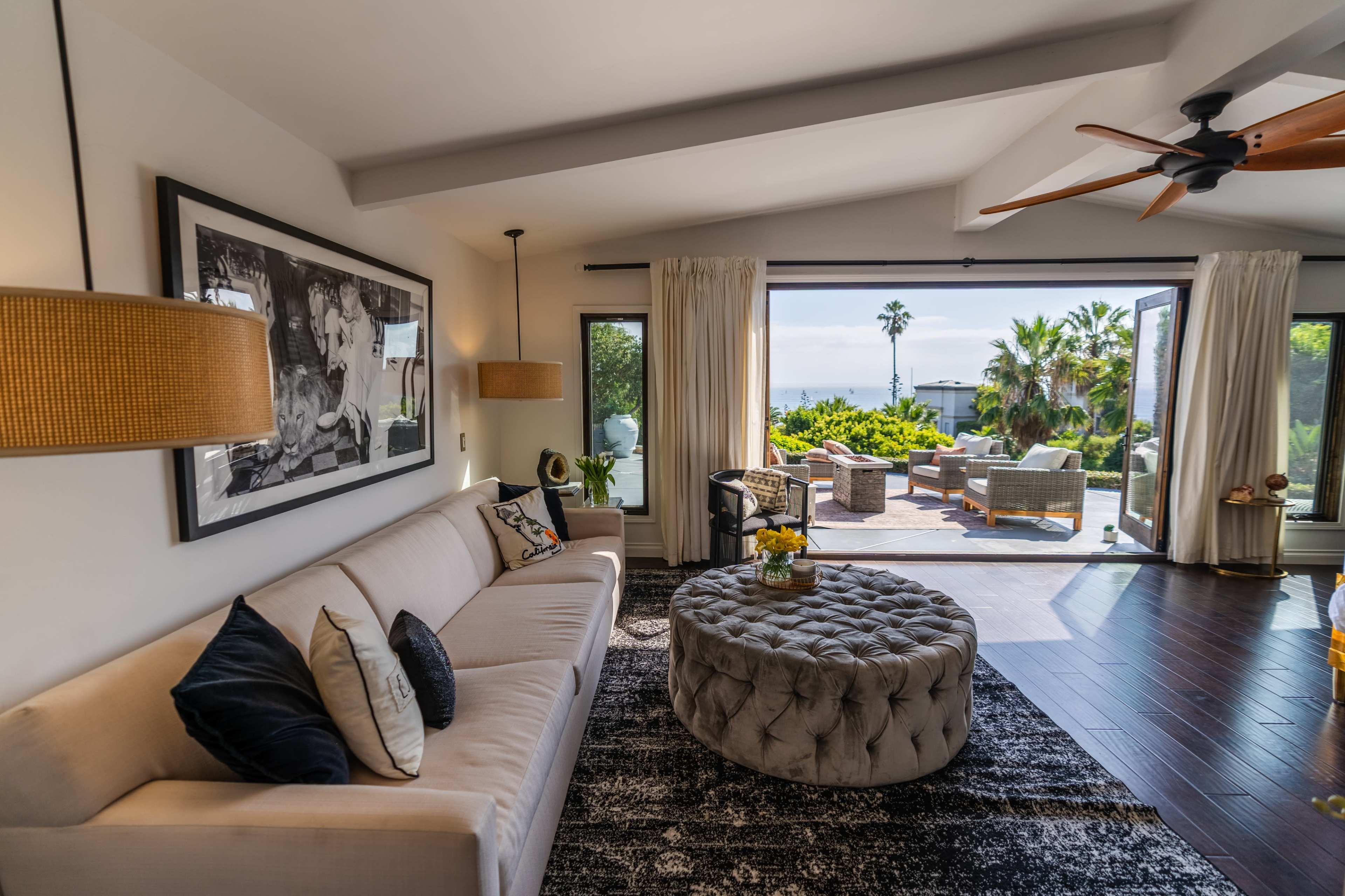 A modern living room featuring a light-colored sofa, a circular tufted ottoman, and large windows opening to an outdoor seating area surrounded by greenery.