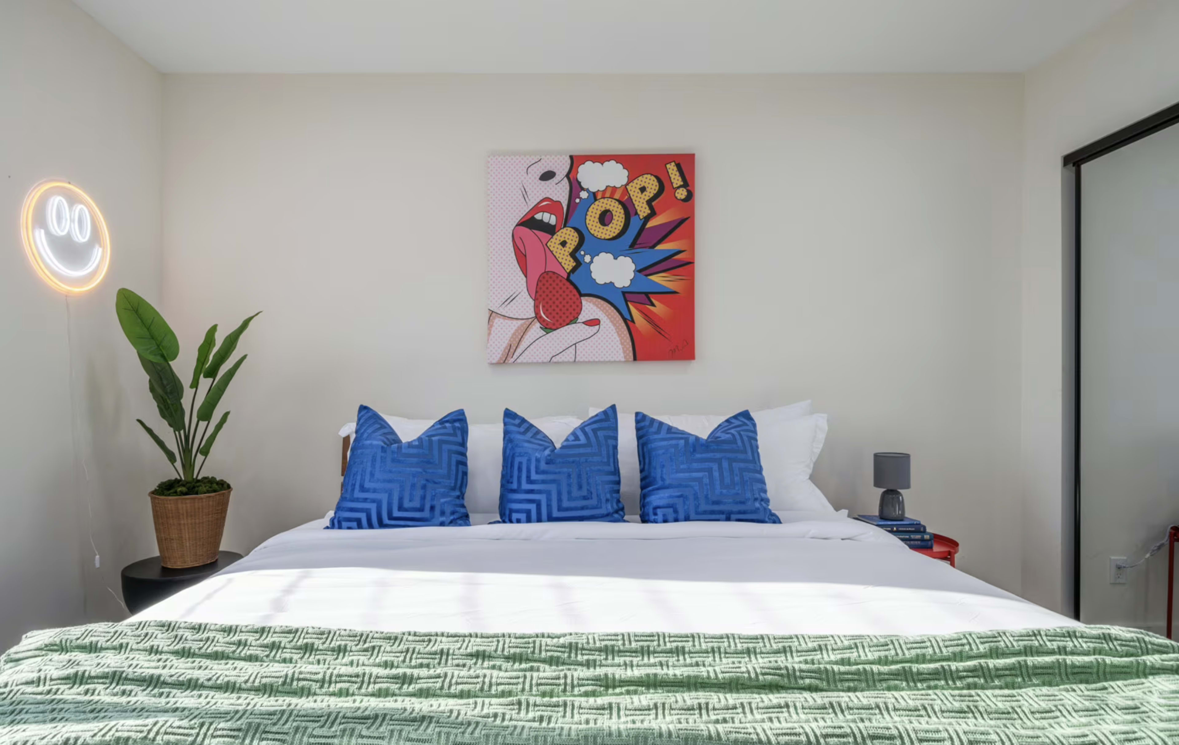 The image shows a modern bedroom featuring a bed with white linens, blue decorative pillows, a green throw blanket, a potted plant, and a colorful pop art piece on the wall.