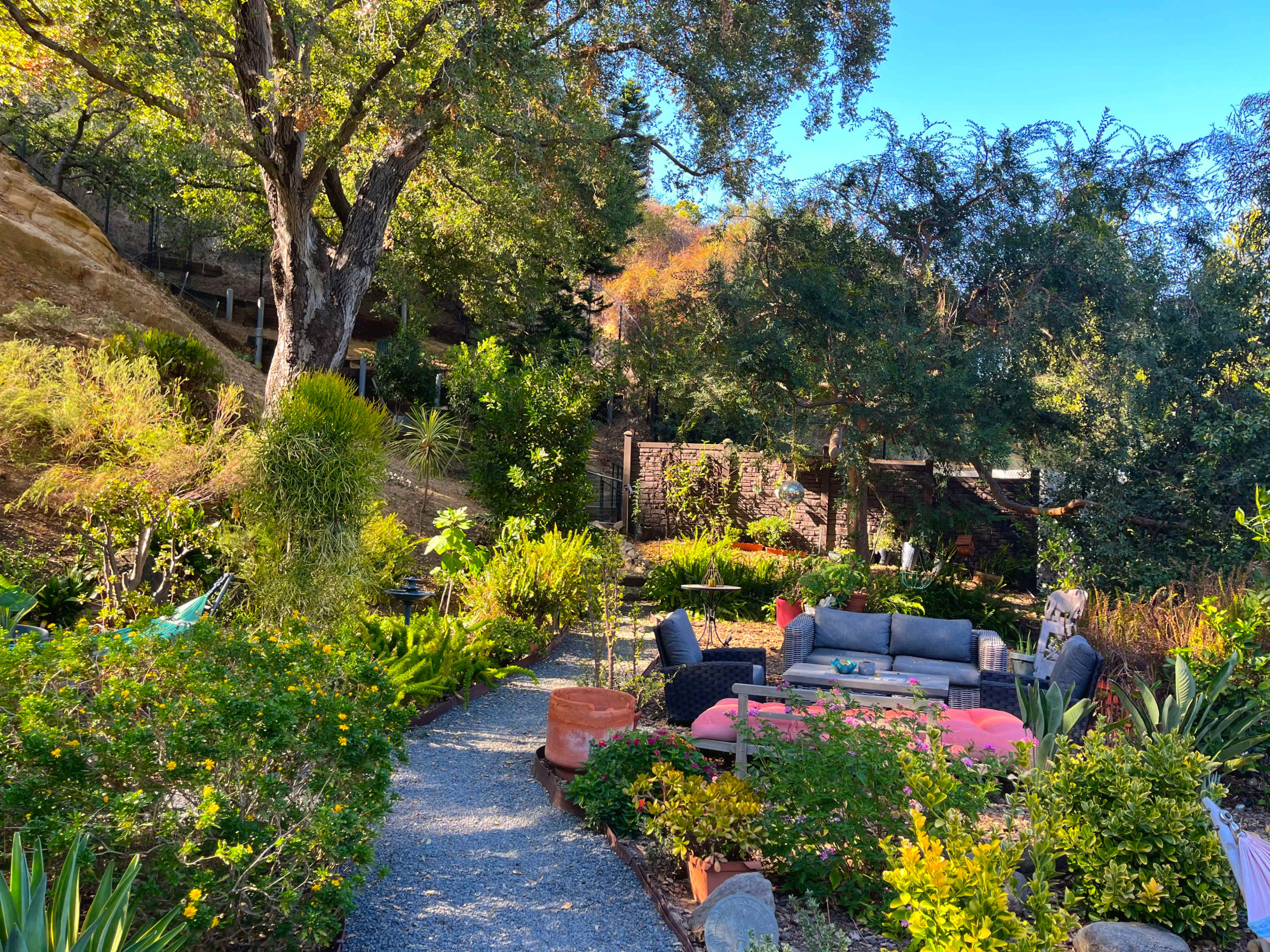 A winding gravel path leads through a vibrant garden filled with various plants and comfortable seating areas under a clear blue sky.