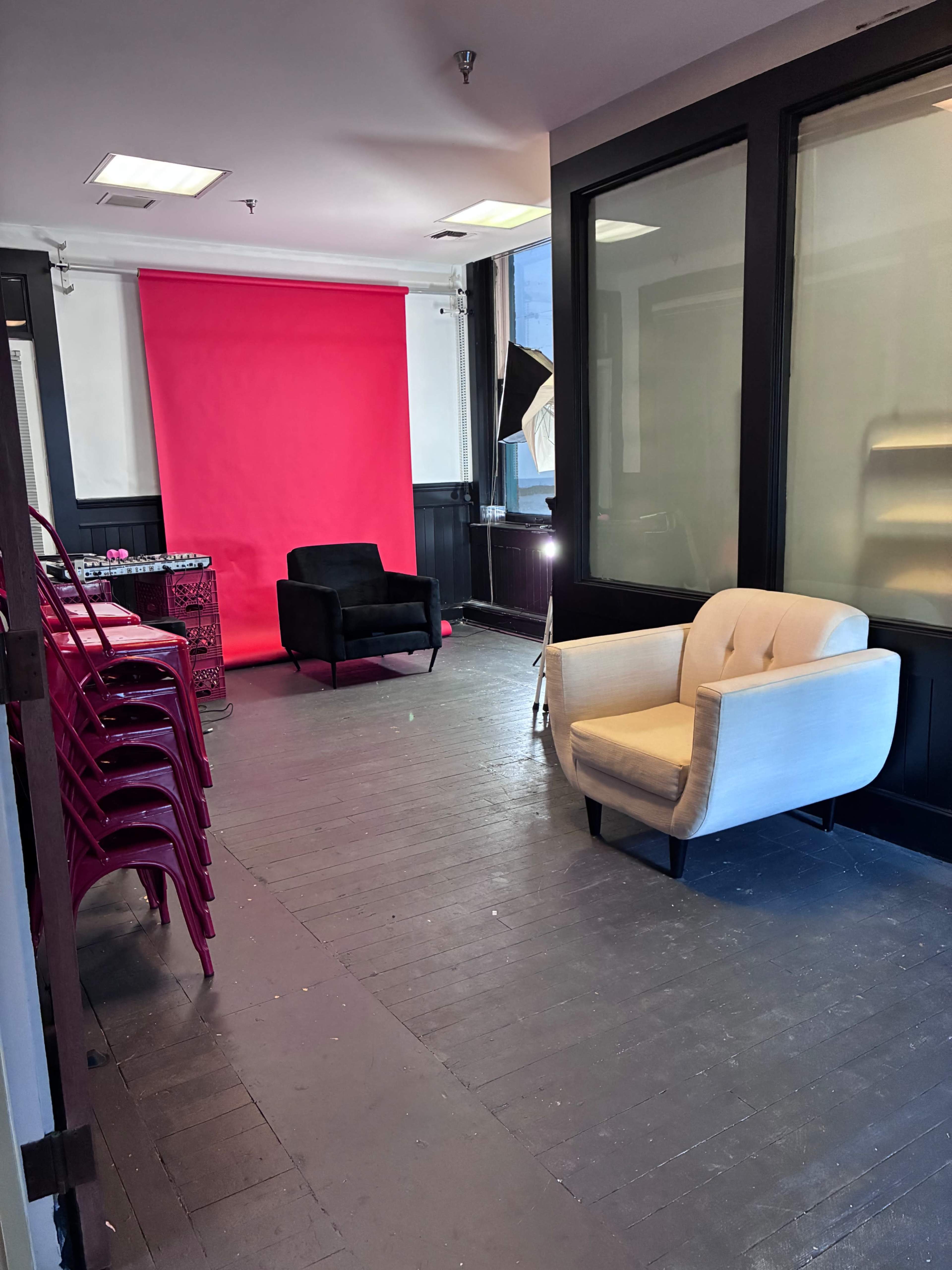 The image shows a room with pink walls, two armchairs, and several stacked chairs, with lighting equipment set up for photography.