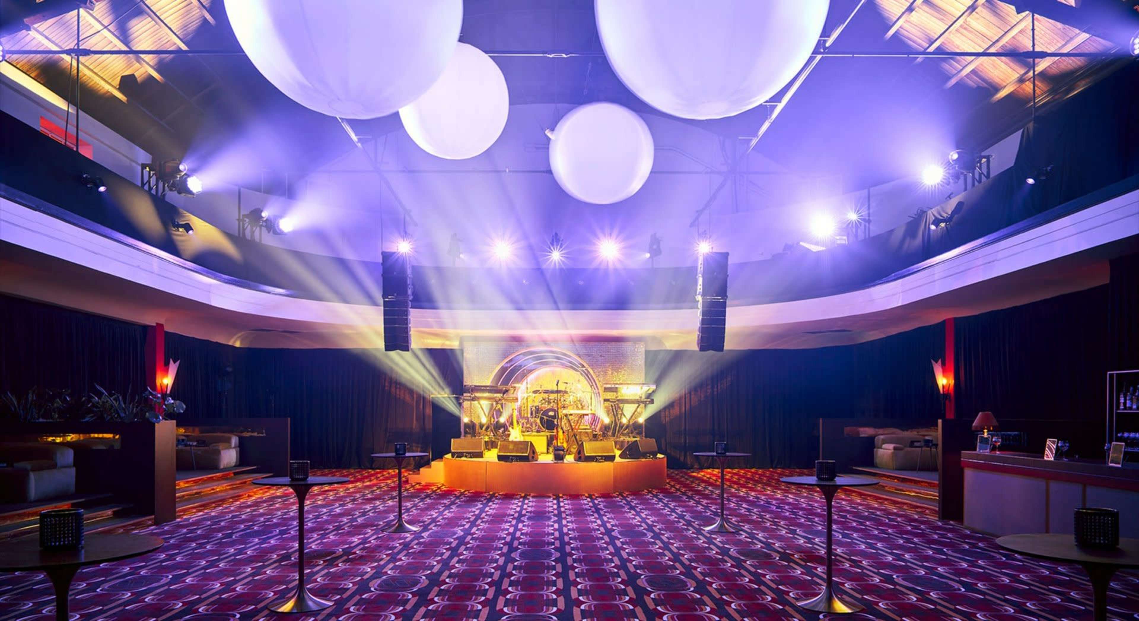 A brightly lit event space features a stage with musical instruments, surrounded by a spacious area with seating and decorative lighting.